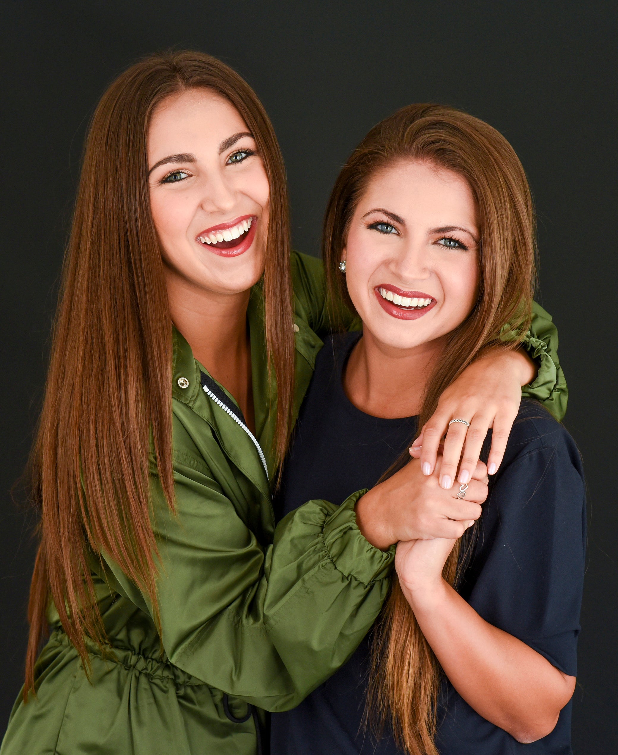 Two women with long hair smiling and embracing each other against a dark background