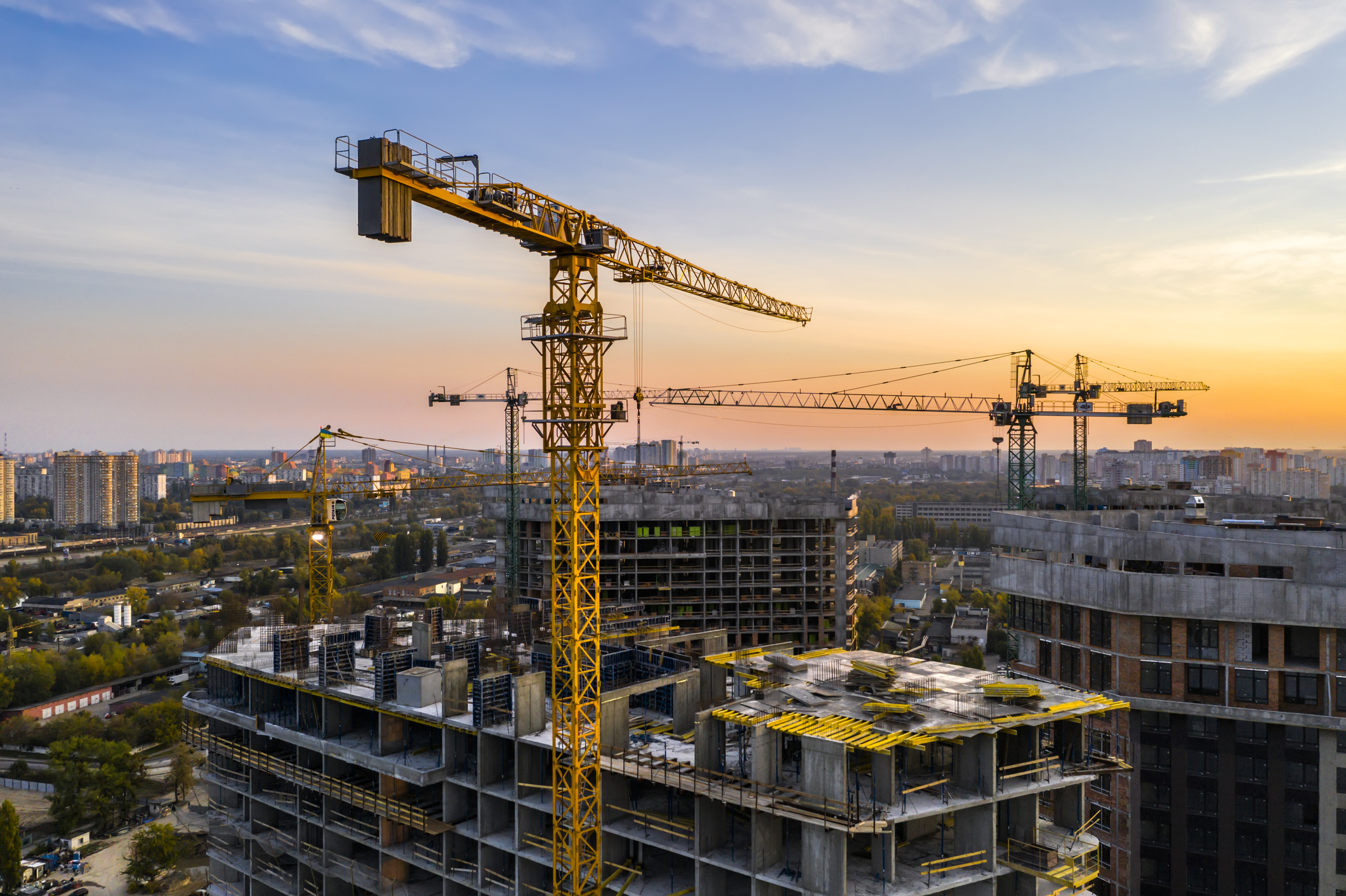 Construction site with multiple cranes working on high-rise buildings, cityscape visible in the background during sunset