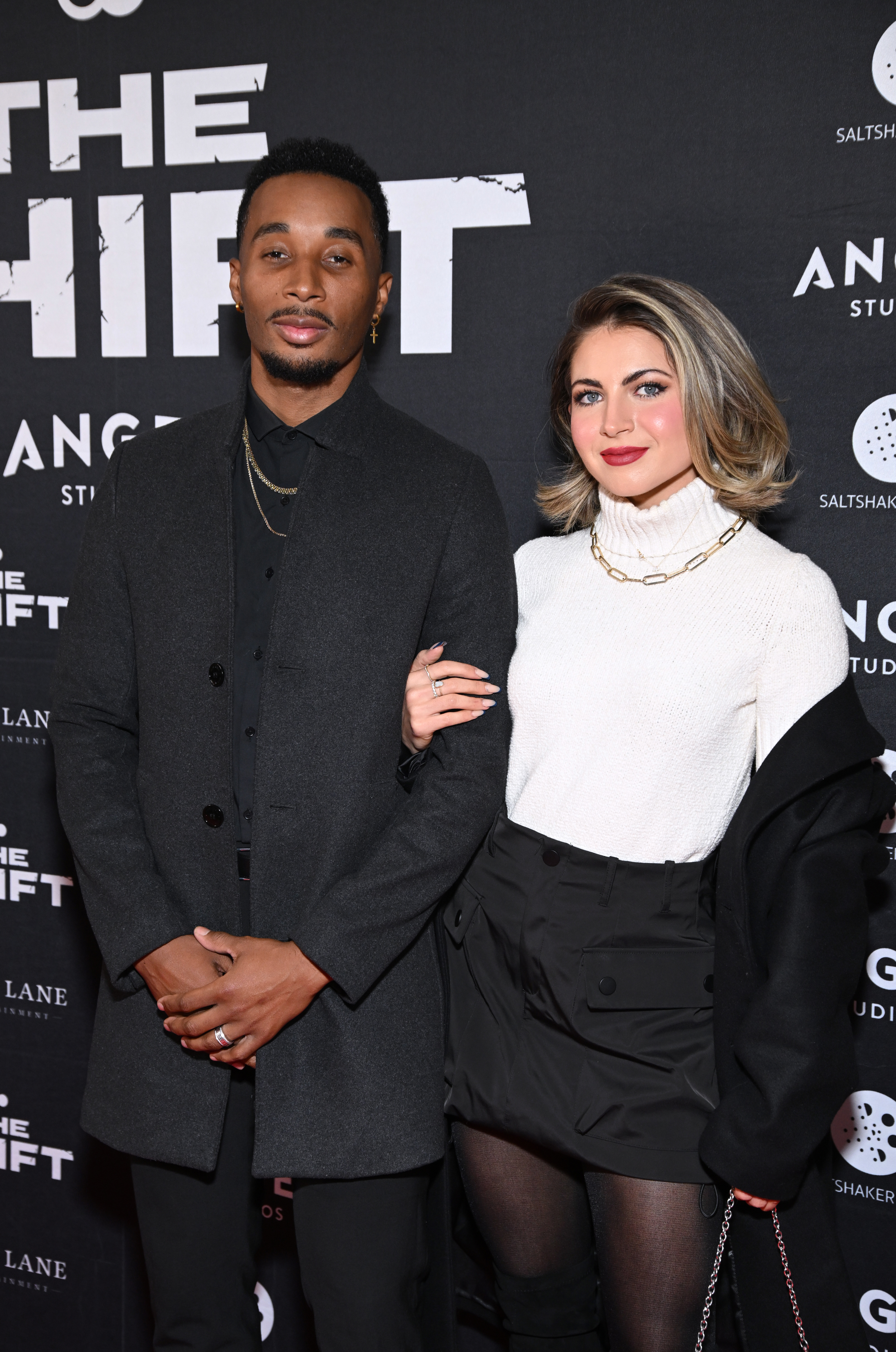 Two individuals posing together at a red carpet event for &quot;The Shift.&quot; The man is wearing a dark coat and the woman is wearing a white sweater and black shorts. Names unknown