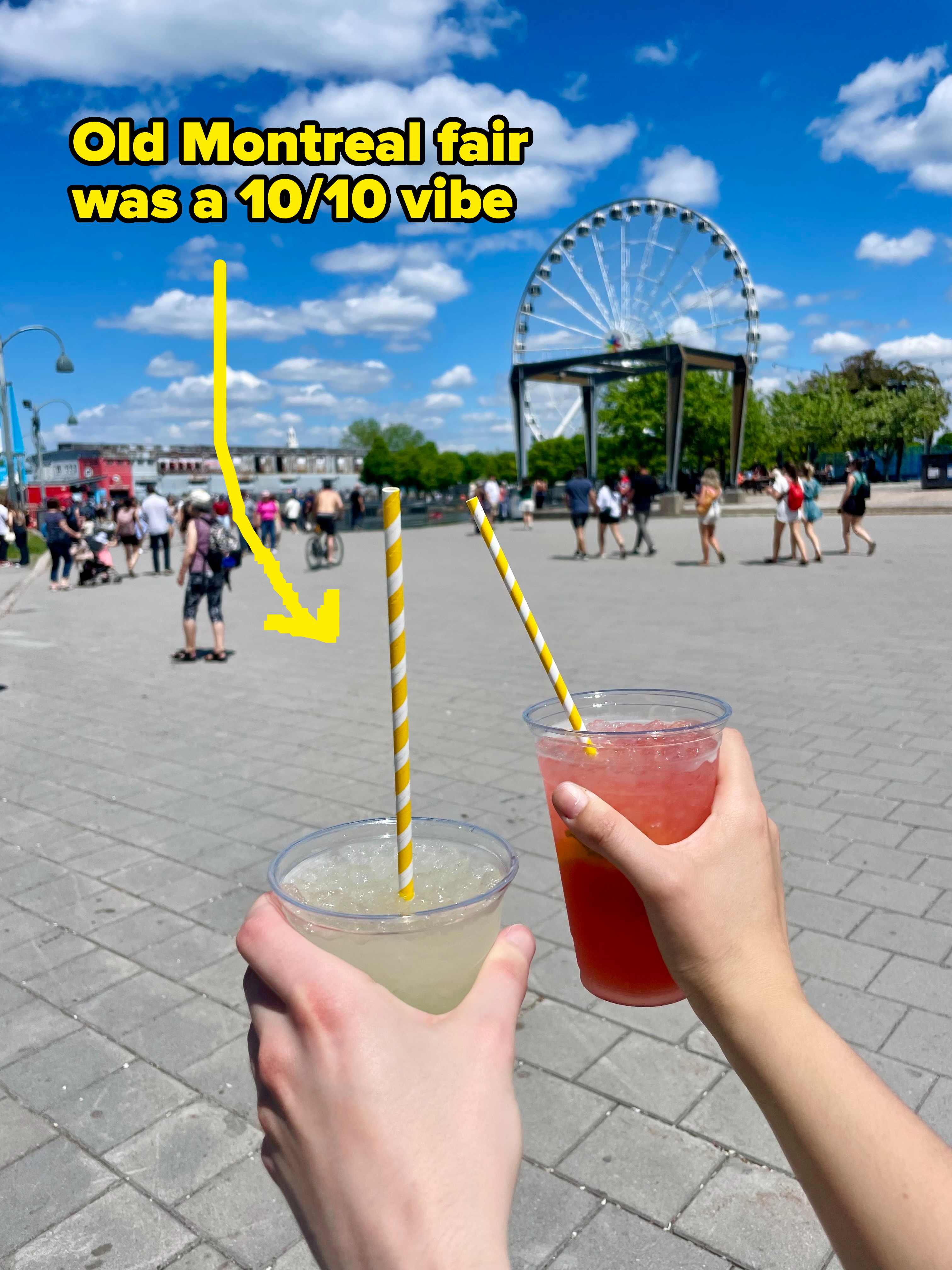 Two hands holding drinks with striped straws in front of a bustling public square with a Ferris wheel in the background
