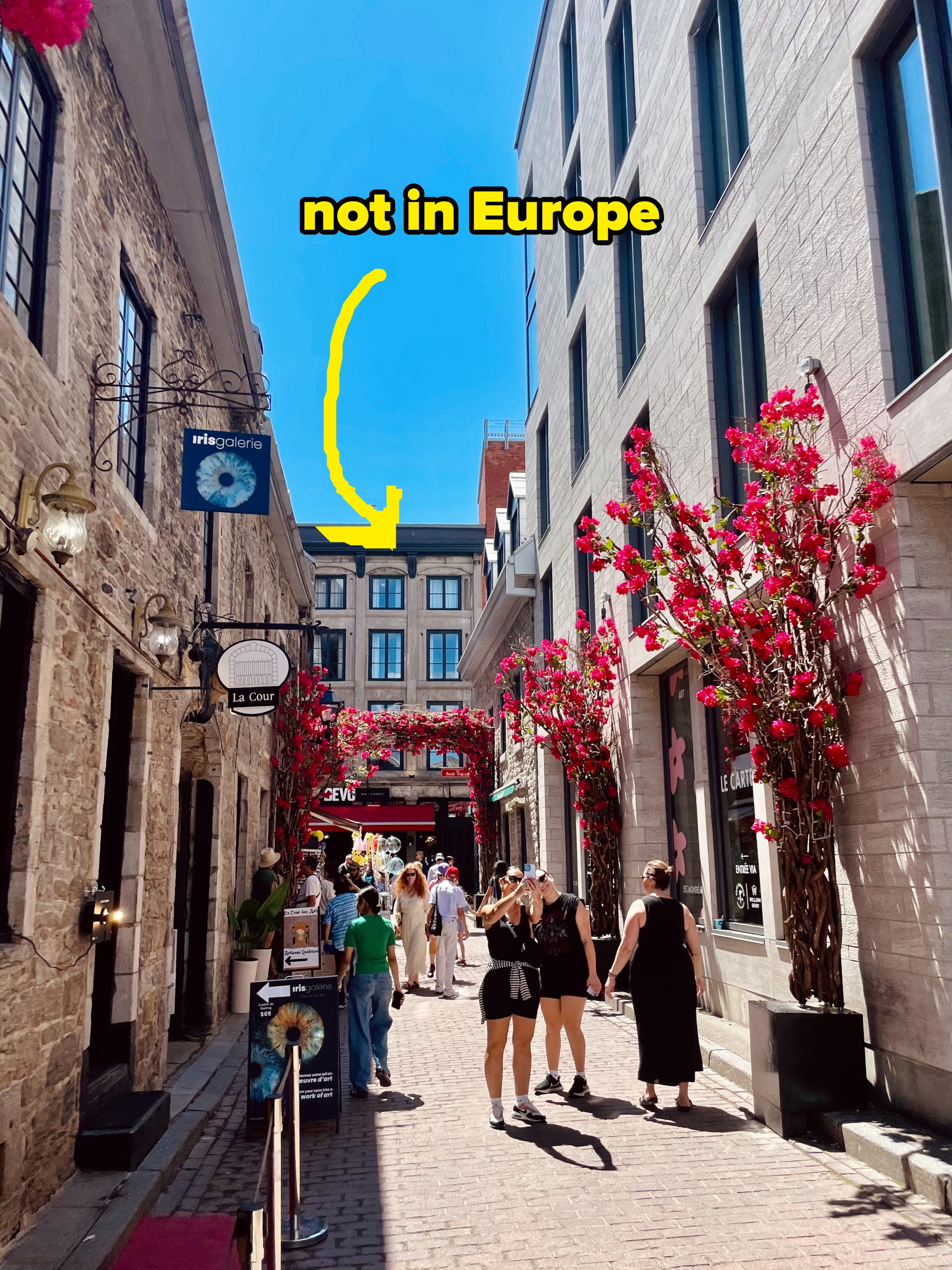 Pedestrians walk down a lively cobblestone street with buildings adorned with red flowers. A large text overlay reads, "not in Europe" with an arrow pointing at the buildings