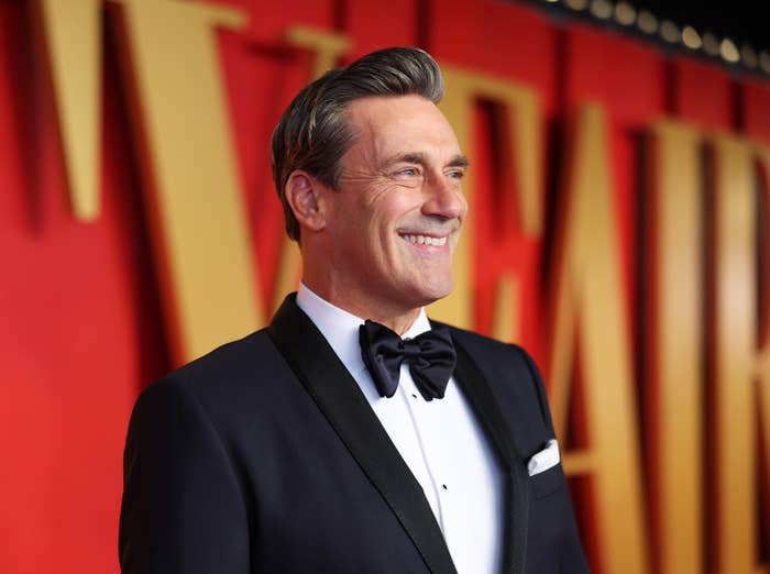 Jon Hamm, in a classic tuxedo with a black bow tie, smiles while standing in front of a "Veep" premiere backdrop
