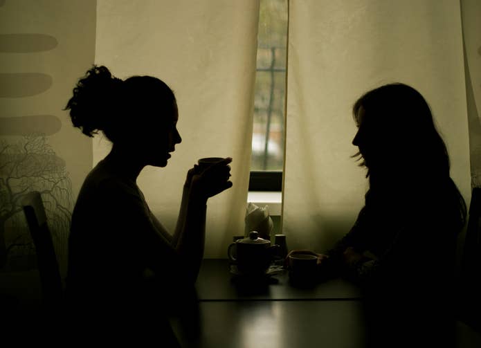 Two people sit at a table, silhouetted against a window, engaging in conversation over coffee. They are facing each other in a dimly lit room