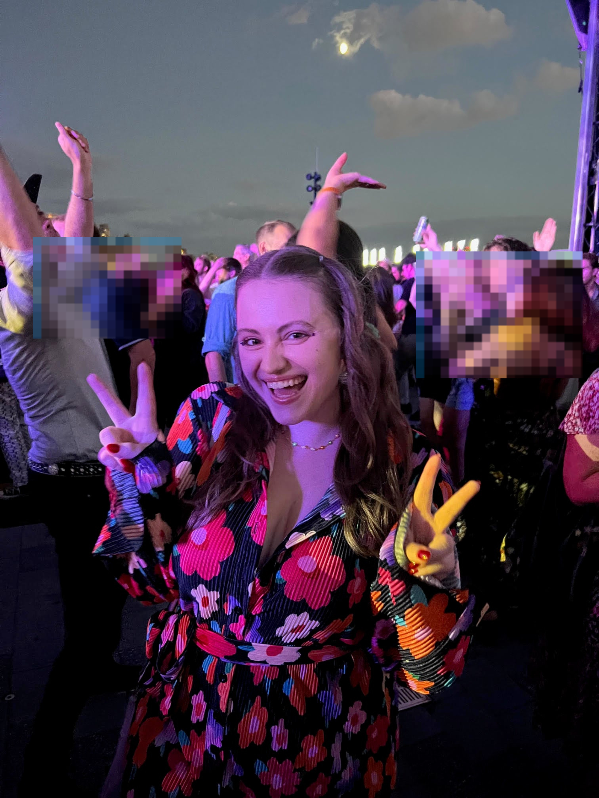 A person in a floral pattern outfit smiles and holds up peace signs at a lively outdoor concert with a crowd dancing and enjoying the performance