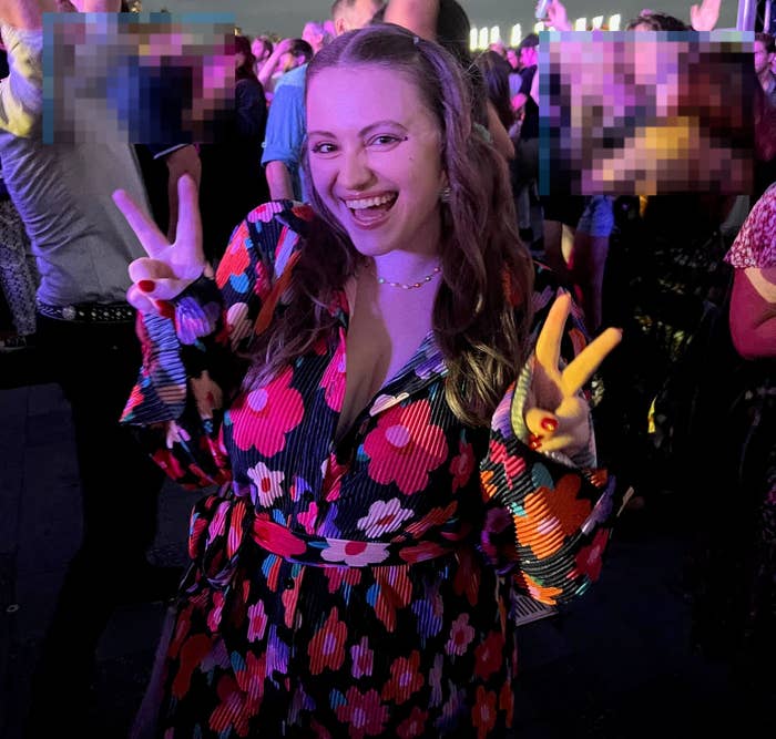 A person in a floral pattern outfit smiles and holds up peace signs at a lively outdoor concert with a crowd dancing and enjoying the performance