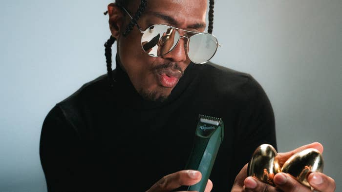 Nick Cannon, wearing a black turtleneck and sunglasses, looks at a clipper held in one hand and two golden eggs in the other