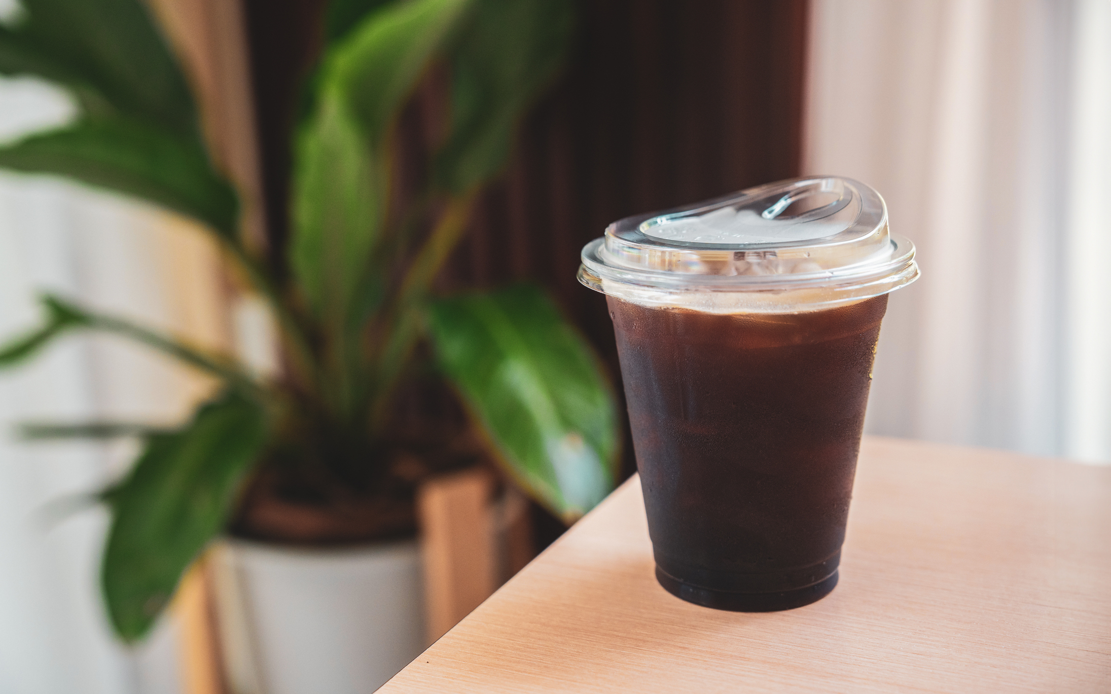 An iced coffee in a plastic cup with a lid sits on a wooden table with a plant in the background