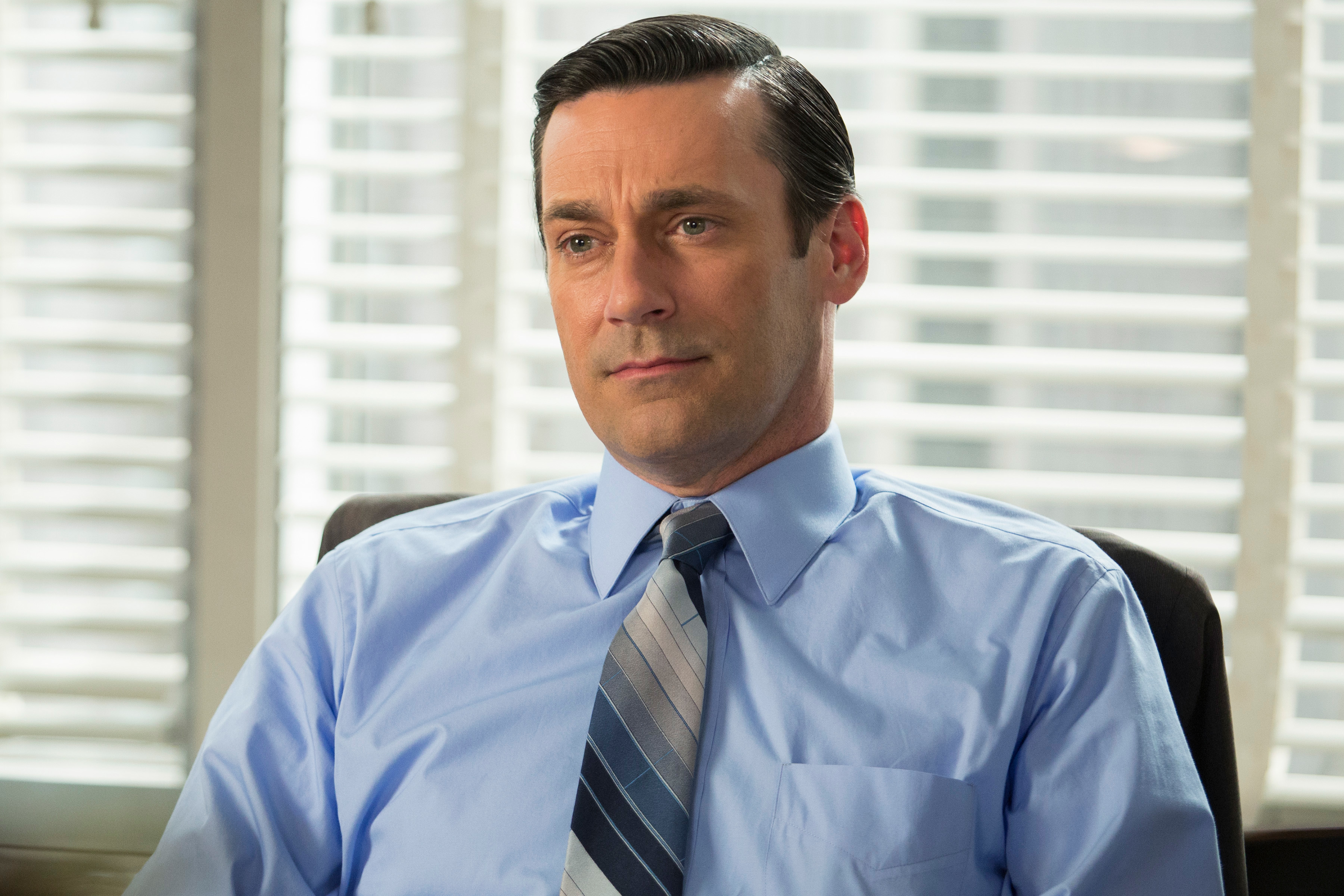 Jon Hamm sits indoors in a formal setting, wearing a light blue dress shirt and striped tie. He has a calm and composed expression