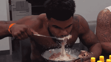 A shirtless man with tattoos eagerly eats a large bowl of cereal, splashing milk around