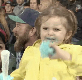 A young girl, standing in a crowd at a sports event, joyfully eats cotton candy, with some on her face