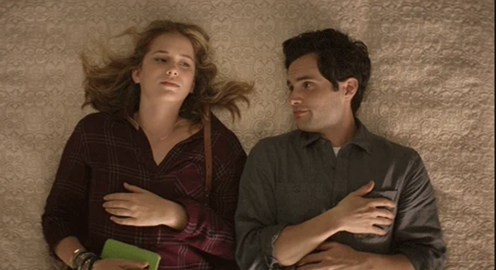 Elizabeth Lail and Penn Badgley lie on a bed, both looking contemplative while staring at each other, with relaxed, casual clothing