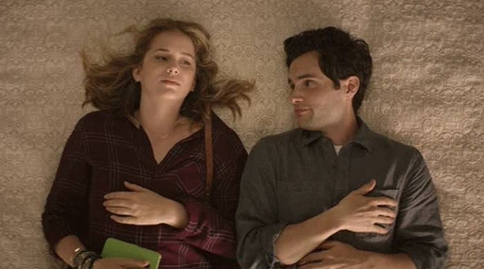 Elizabeth Lail and Penn Badgley lie on a bed, both looking contemplative while staring at each other, with relaxed, casual clothing