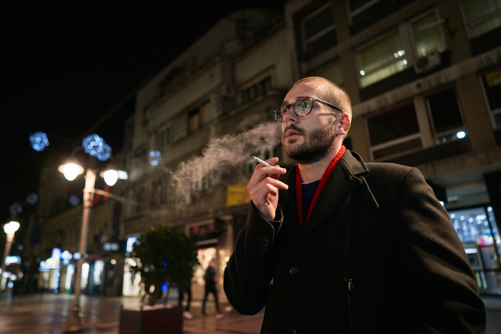 A bearded man in glasses and a black coat smokes a cigarette on a city street at night