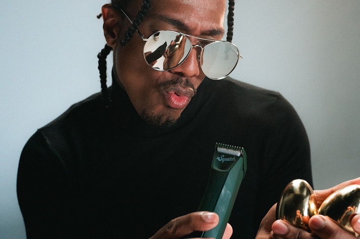 Nick Cannon, wearing a black turtleneck and sunglasses, looks at a clipper held in one hand and two golden eggs in the other