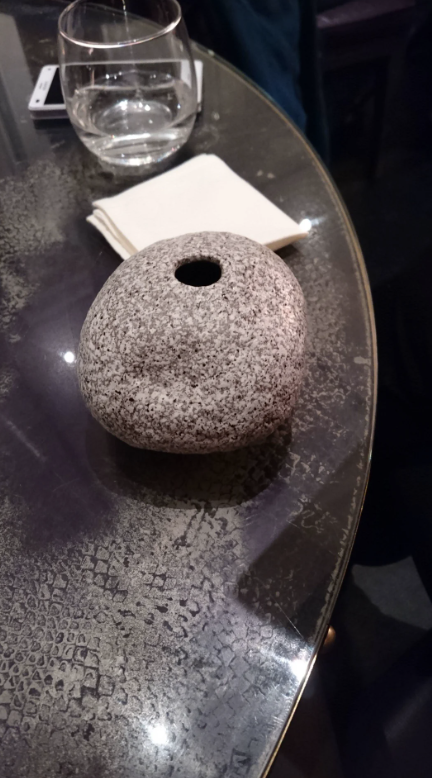 A stone with a hole, resembling a natural sculpture, on a textured tabletop beside a glass and a paper napkin