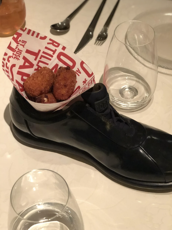 A unique food presentation features fried croquettes served in a black dress shoe on a formal dining table with glassware and utensils
