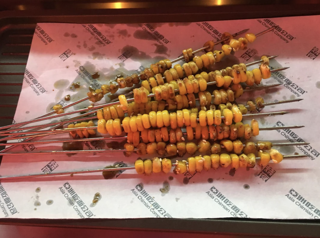 Skewered corn kernels are laid out on a food tray, likely from a market or food festival, resting on grease-stained parchment paper