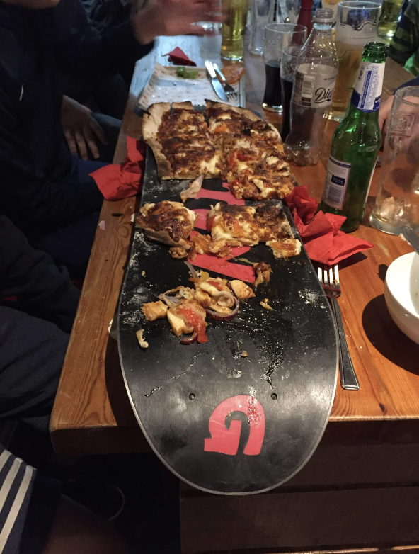 A group of people are seated at a table, sharing a pizza served on a snowboard. Several half-eaten slices are visible with drinks around