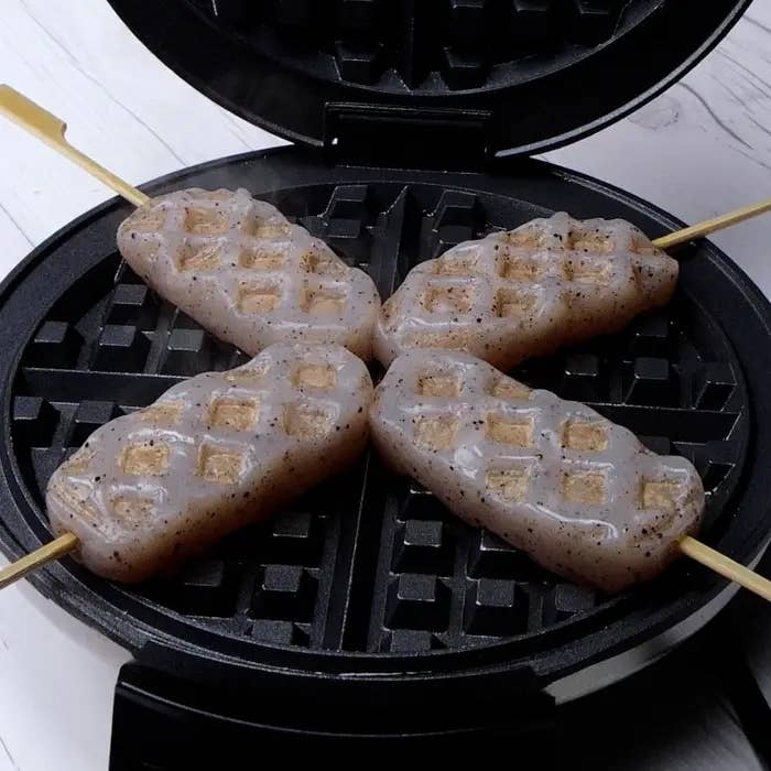 ワッフルメーカーで焼かれている4本の棒に刺さった食材。