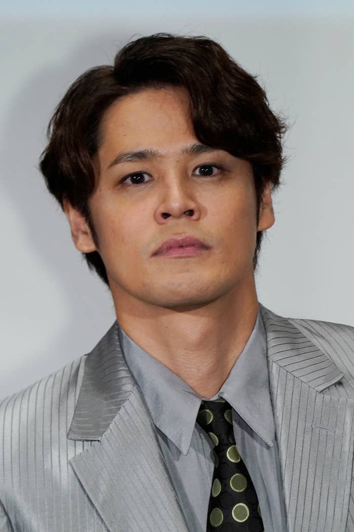 男性がグレーのスーツとドット柄のネクタイを着ている。彼はカメラに真剣な表情で見つめている。