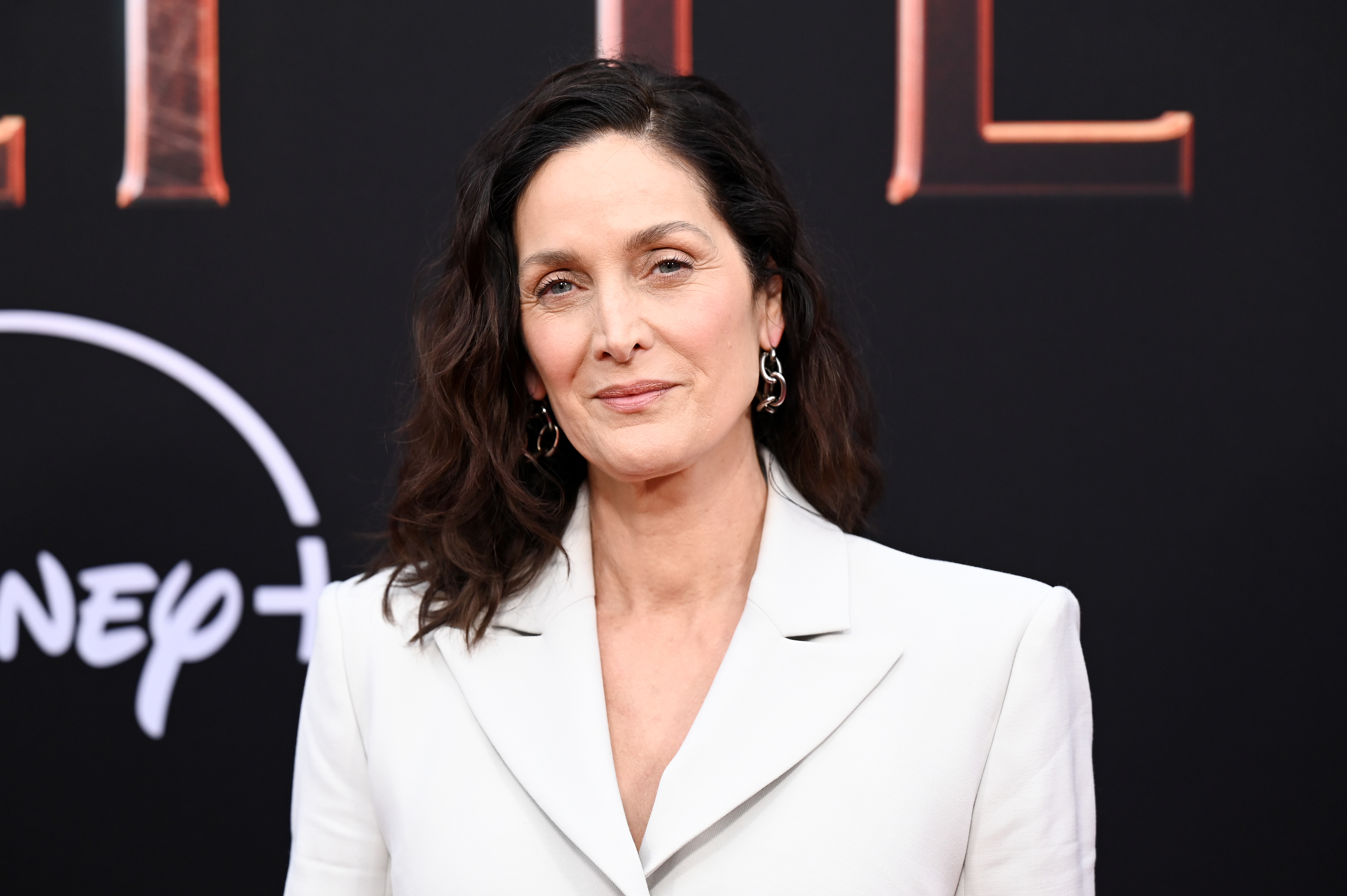 Carrie-Anne Moss at a Disney+ red carpet event, wearing a stylish white blazer