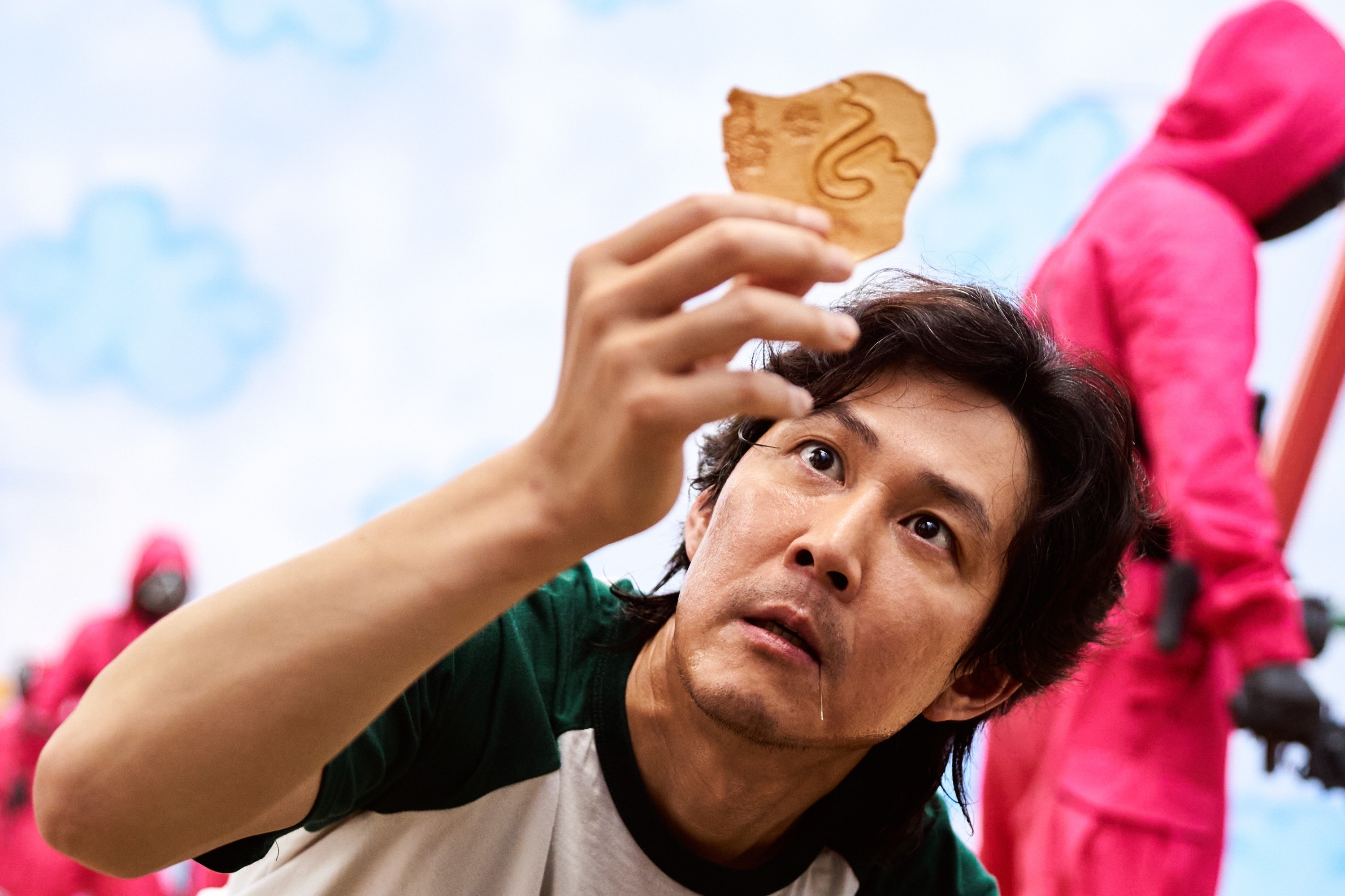 Lee Jung-jae examines a piece in a tense scene from &quot;Squid Game,&quot; with masked guards in red outfits in the background