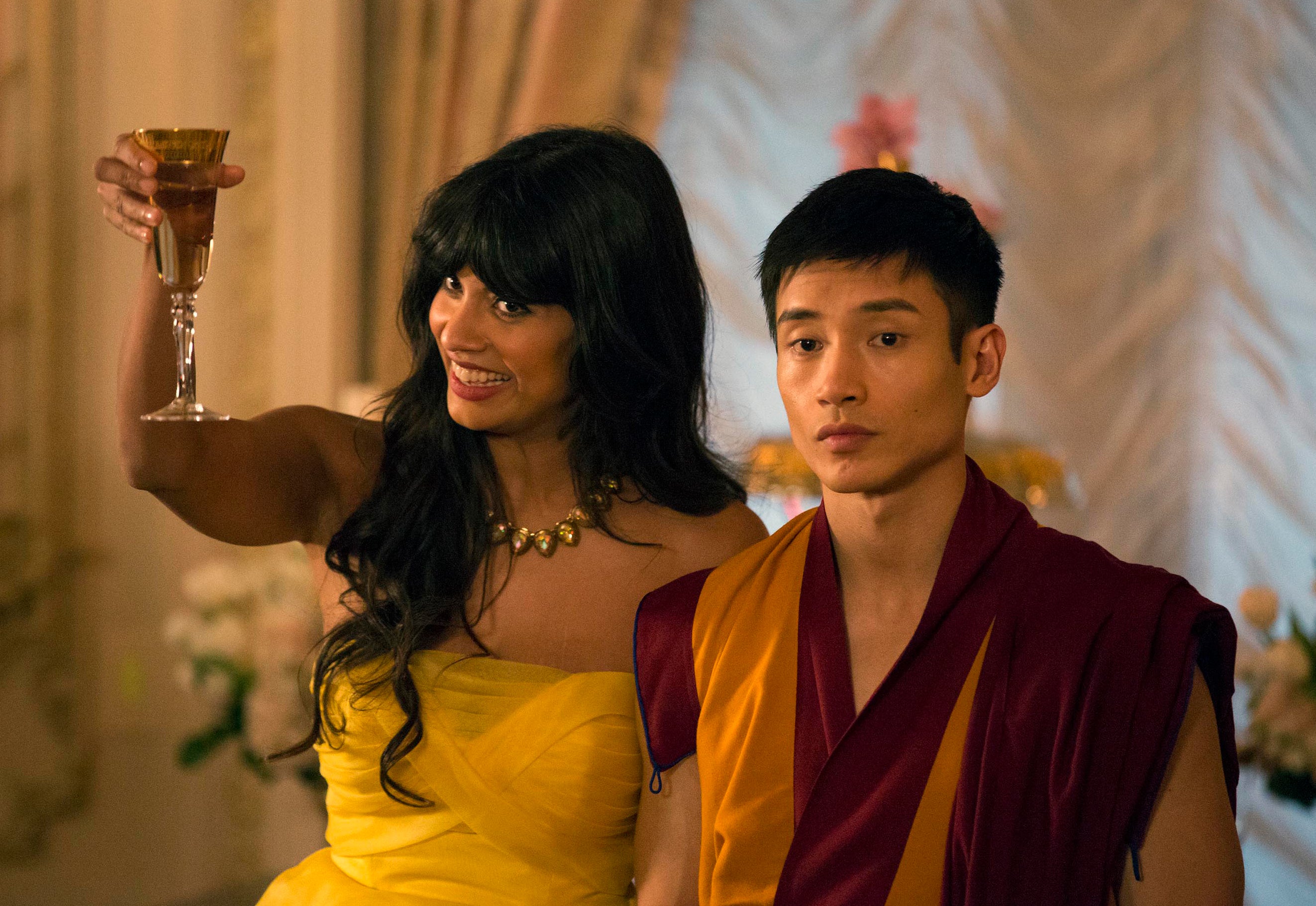 Jameela Jamil raises a glass in a celebratory toast wearing a strapless dress while Manny Jacinto, dressed in a monk robe, stands beside her looking serious