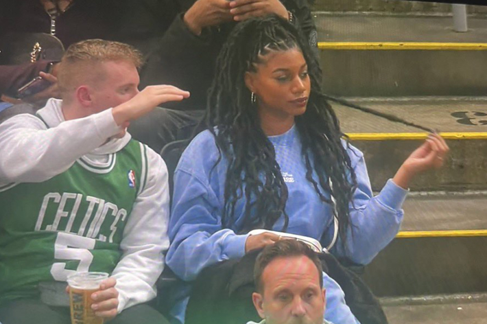 A man wearing a Celtics jersey and holding a drink sits next to a woman in a blue sweater with long braided hair as they watch a sports event