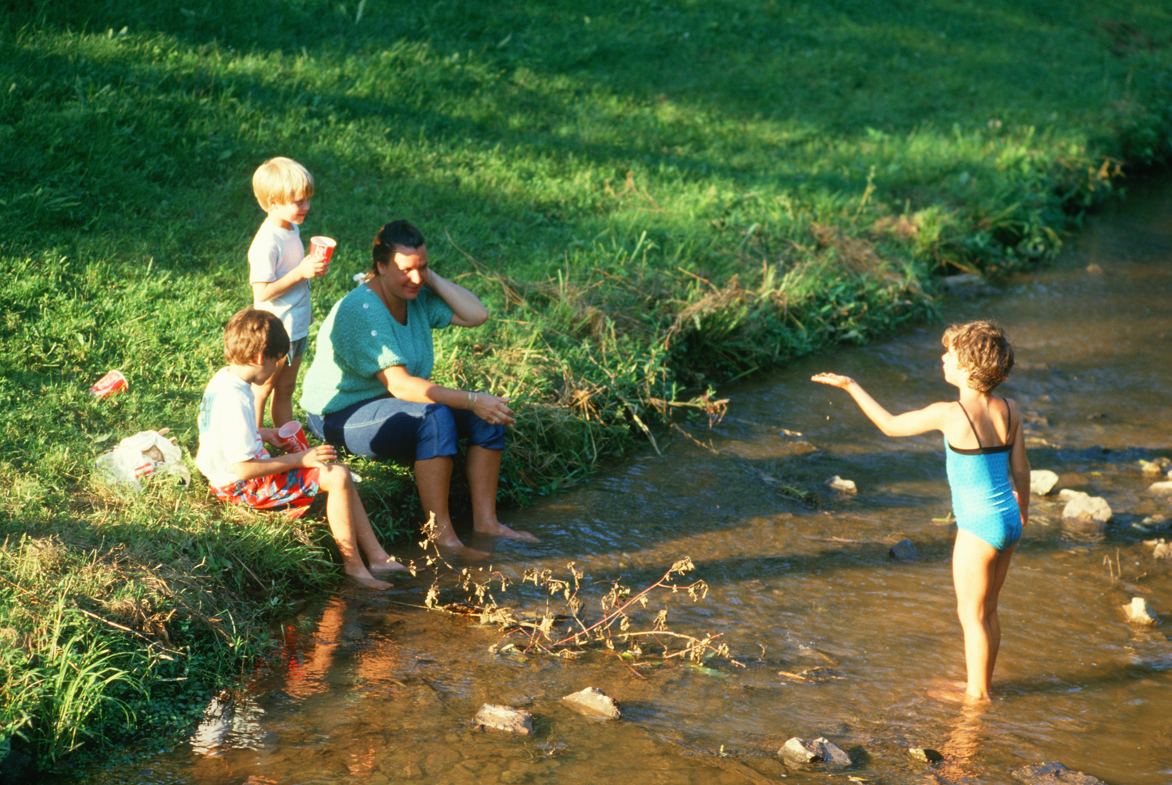 Two adults and two children are near a stream. One child is standing in the water wearing a swimsuit, talking to the others who are seated on the bank