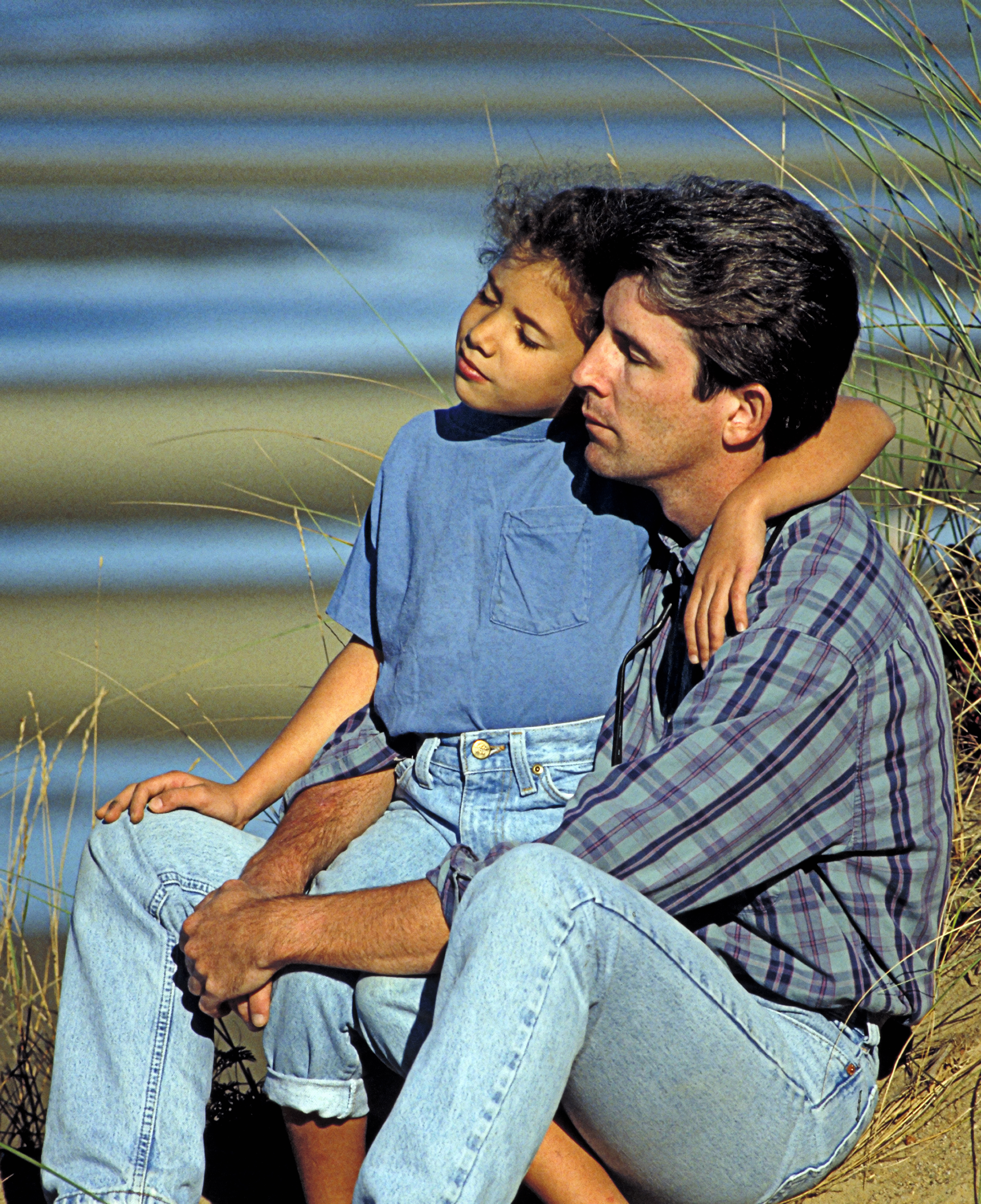 A man and child sit closely together on a beach, both appearing thoughtful. The child is resting their head on the man's shoulder