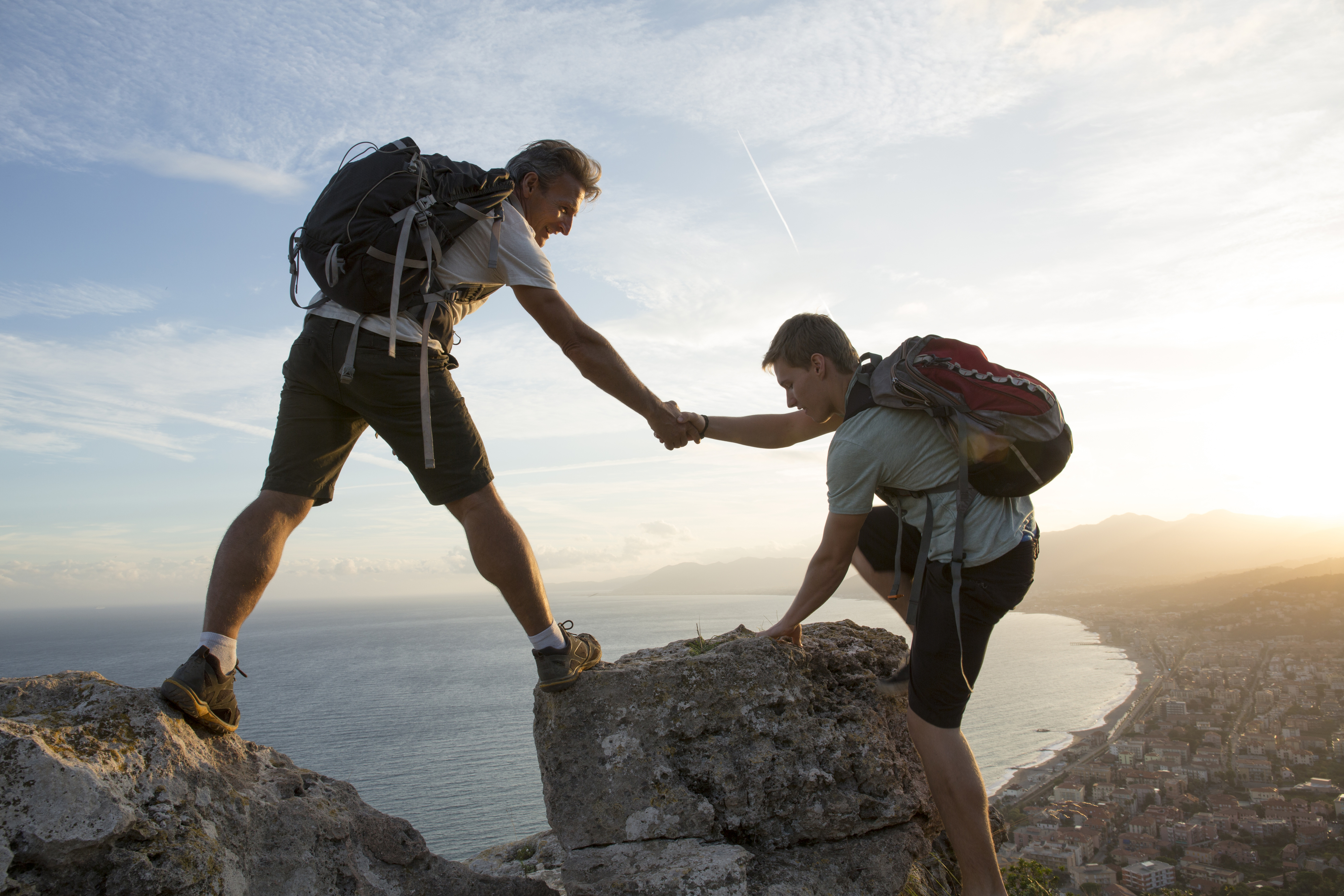 Two hikers assist each other atop a rocky cliff, overlooking a coastal town. Both wear backpacks and outdoor attire