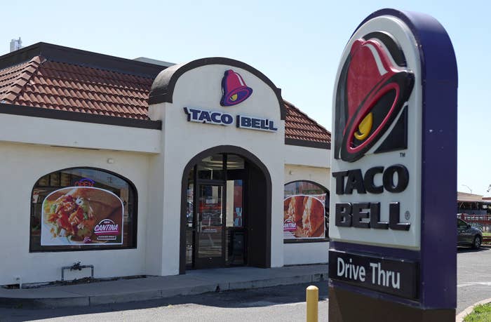 Taco Bell restaurant exterior with a drive-thru sign in the foreground. Large food images are displayed on the restaurant windows