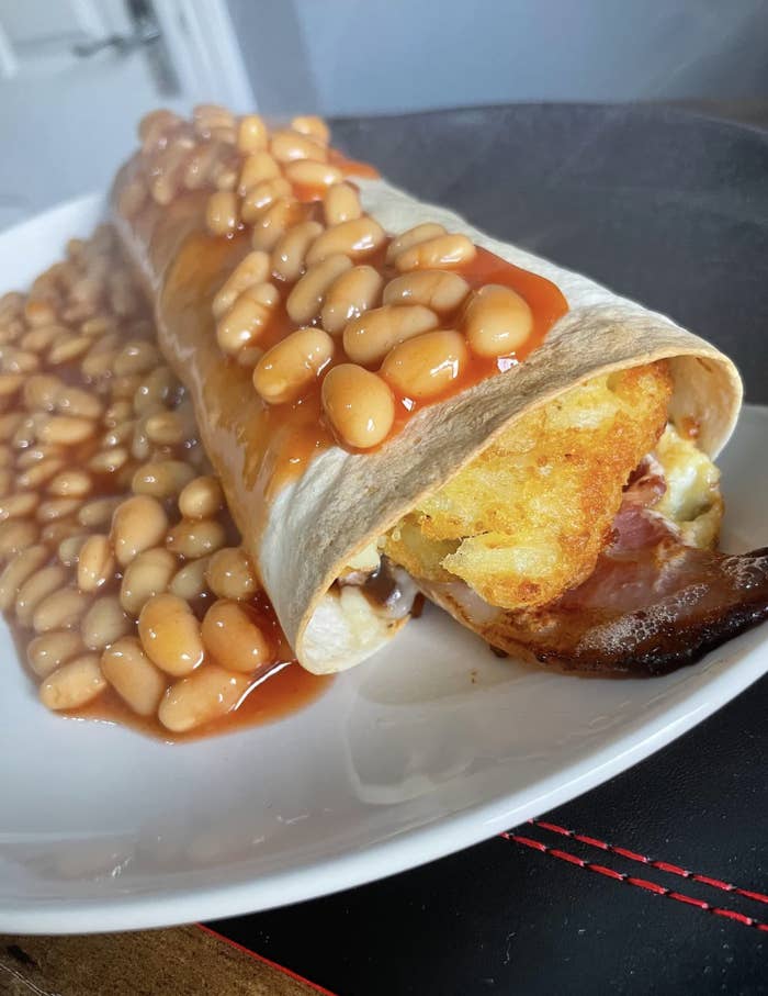 A tortilla wrap filled with egg, bacon, and hash brown topped with baked beans in tomato sauce on a white plate