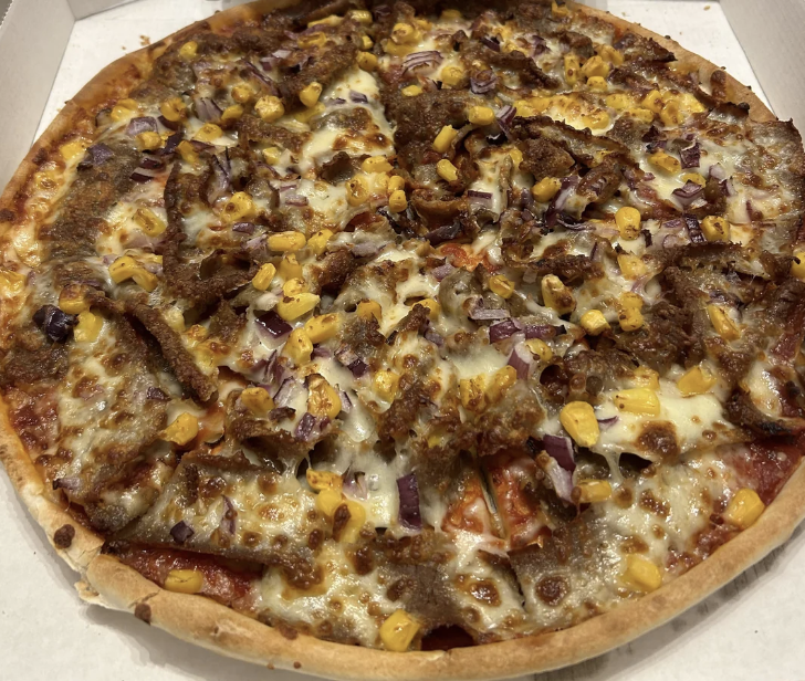 Close-up of a cooked pizza topped with beef, corn, onions, and melted cheese in a cardboard box