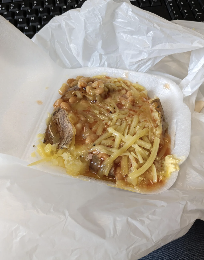 A styrofoam container with cooked meal including beans, shredded cheese, beef, eggplant, and mashed potatoes, placed on a plastic bag. Keyboard in the background