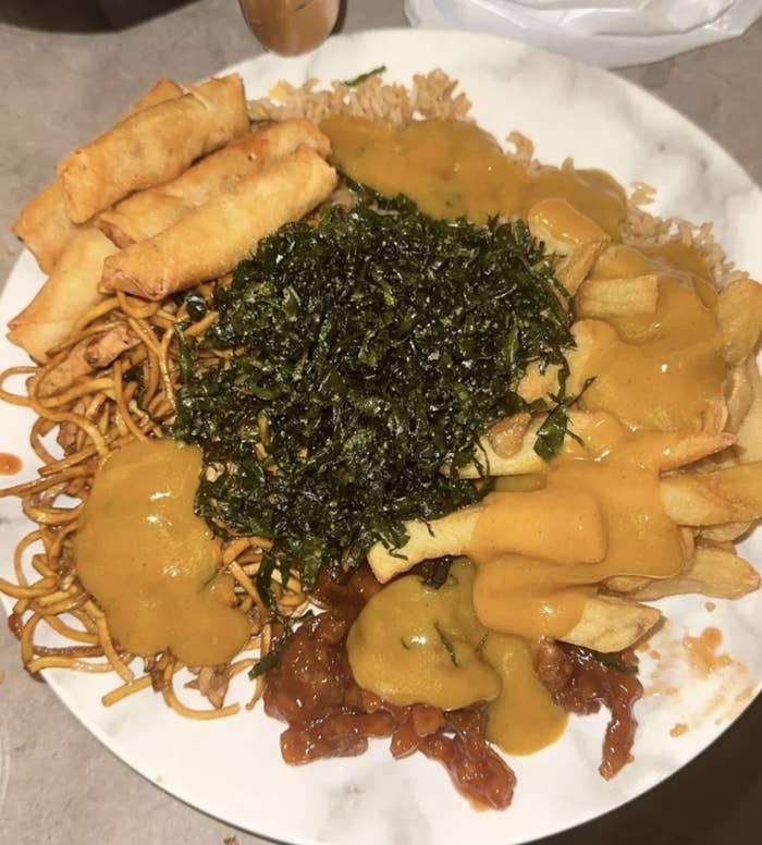 A plate filled with spring rolls, noodles, seaweed, fries, and meat topped with a yellow sauce