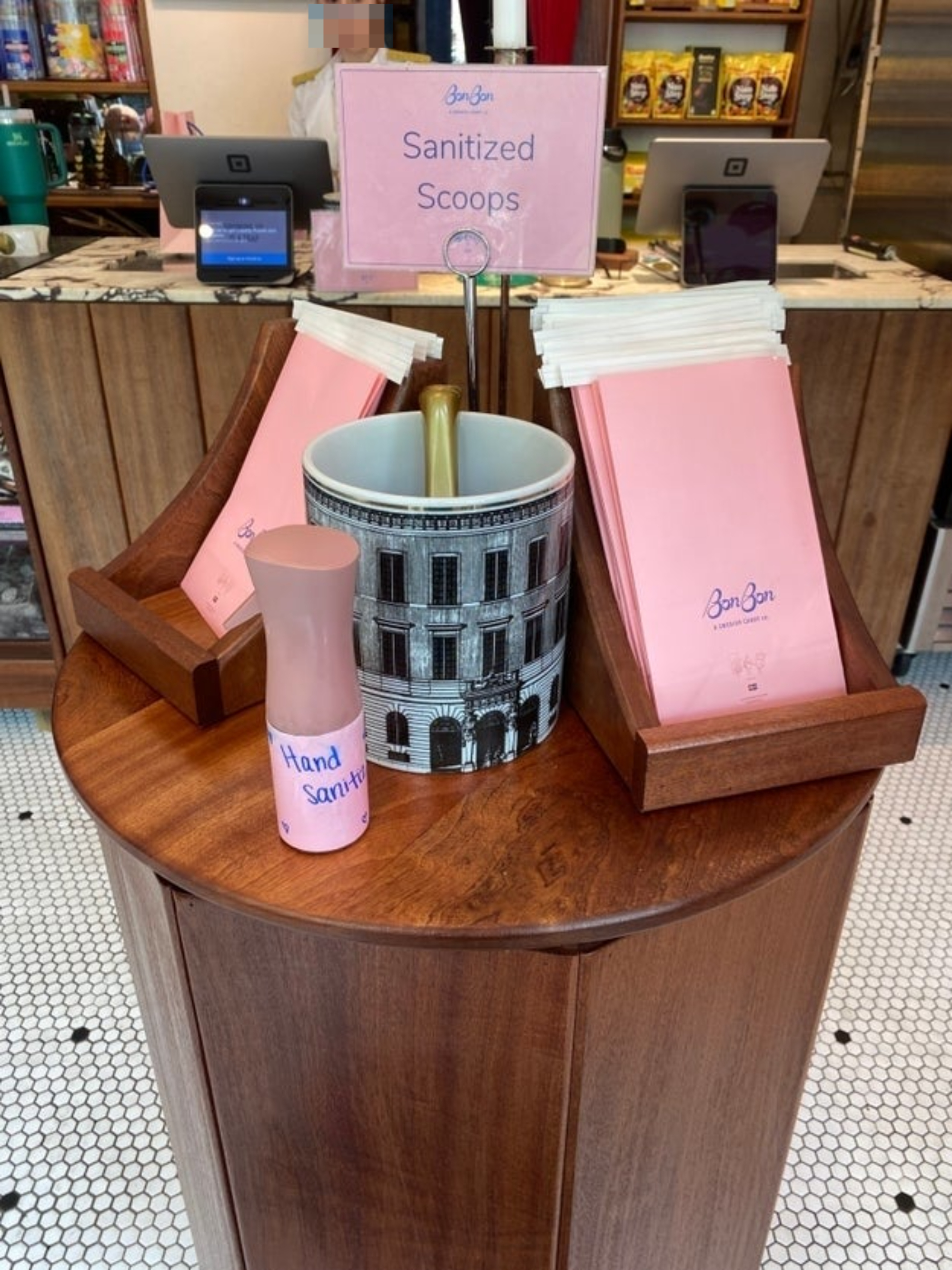 A wooden stand in a shop displaying sanitized scoops, pink napkins, and a bottle of hand sanitizer with a sign saying &quot;BonBon Sanitized Scoops&quot;