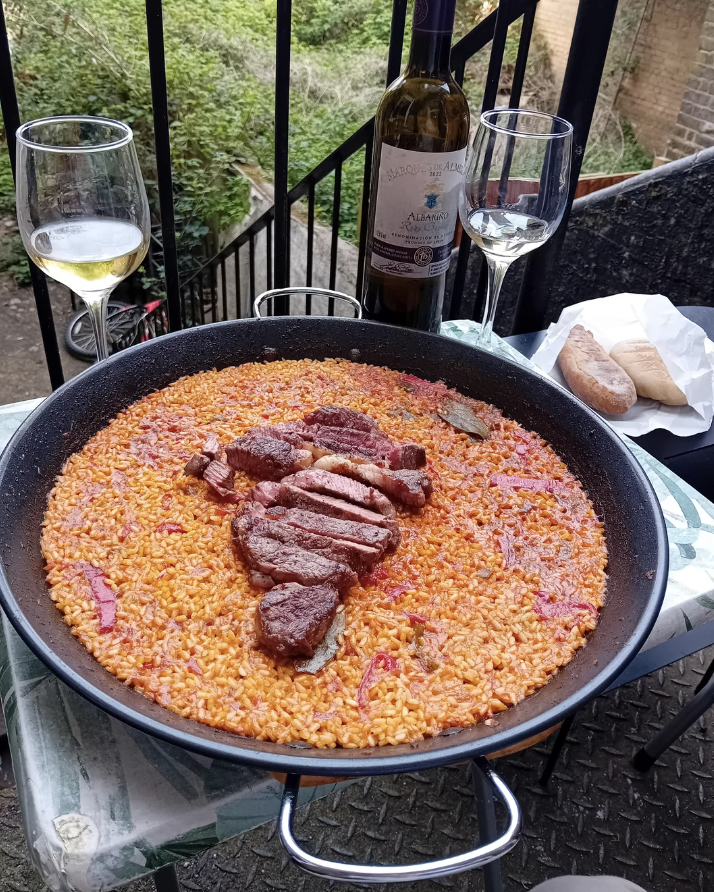 A large pan of paella topped with slices of grilled meat is set on a small outdoor table alongside two glasses of wine, a bottle of wine, and a baguette