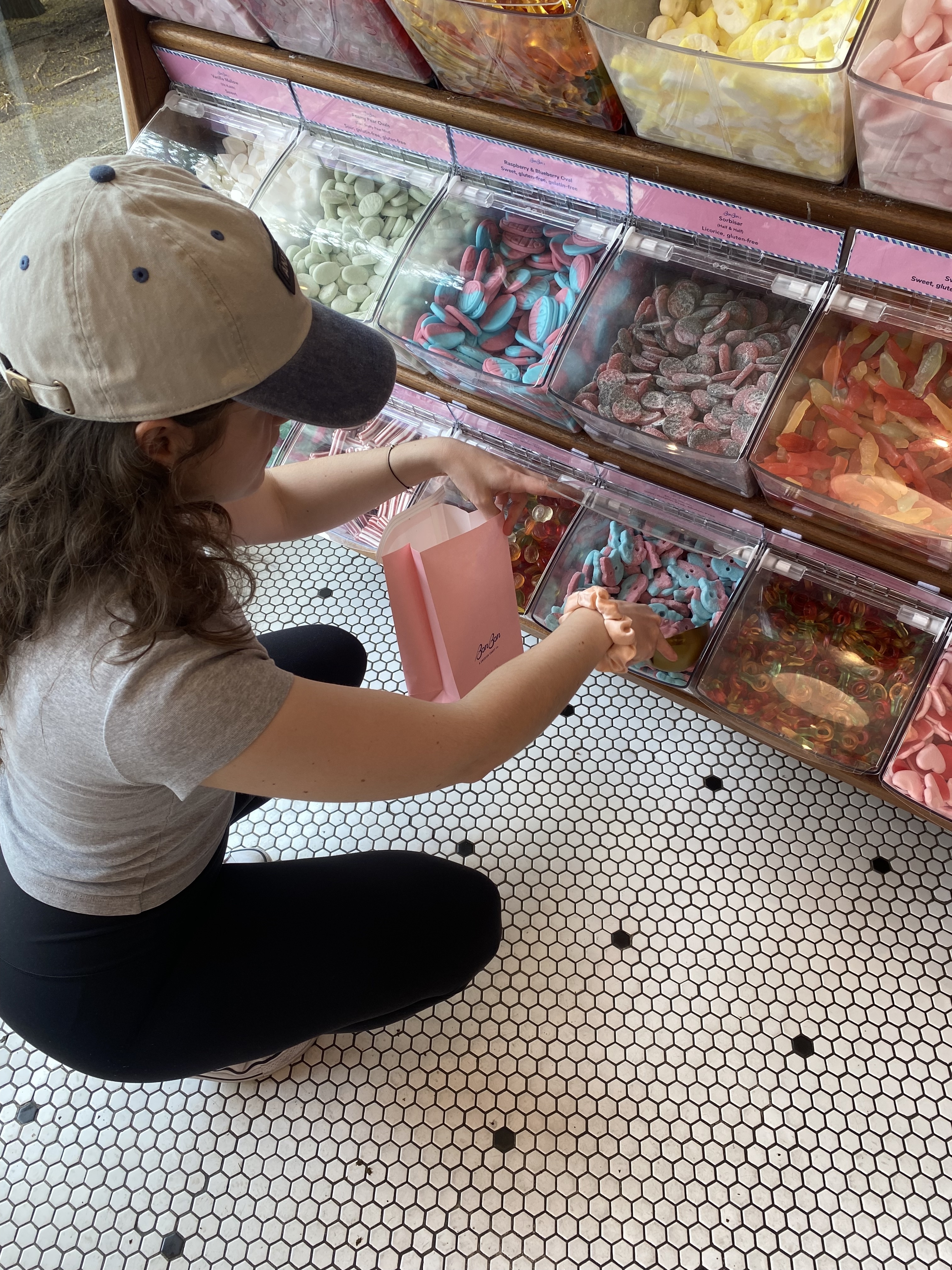 The author in casual clothing fills a pink paper bag with candy from a selection of bins at a candy store