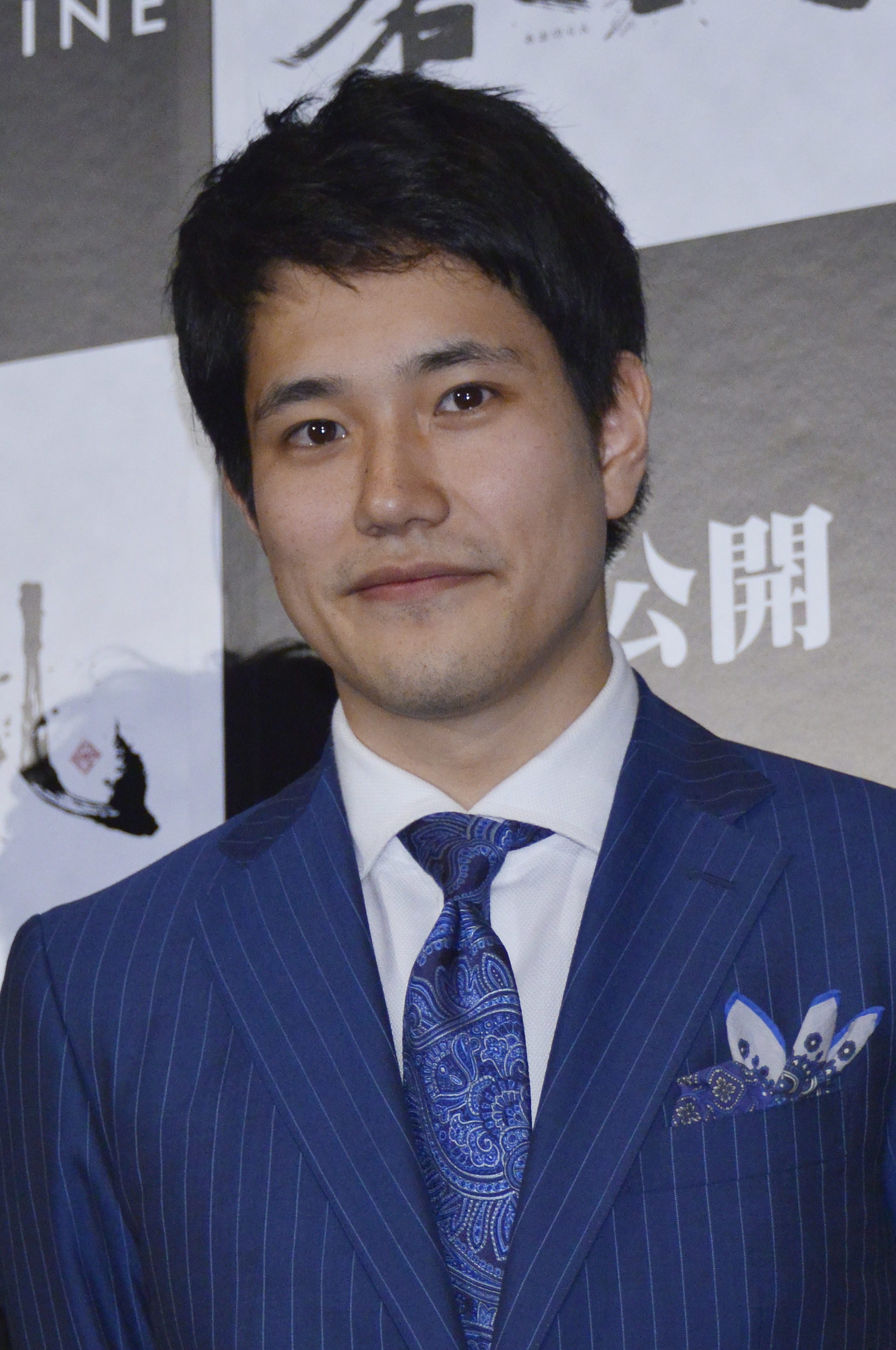 日本の俳優がストライプスーツと装飾されたネクタイを着て微笑んでいる。背景には映画のプロモーション用の看板がある。
