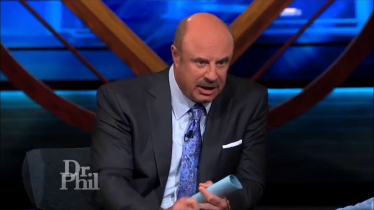 Dr. Phil sits in a dark room, holding papers, appearing serious, likely discussing a topic on his show