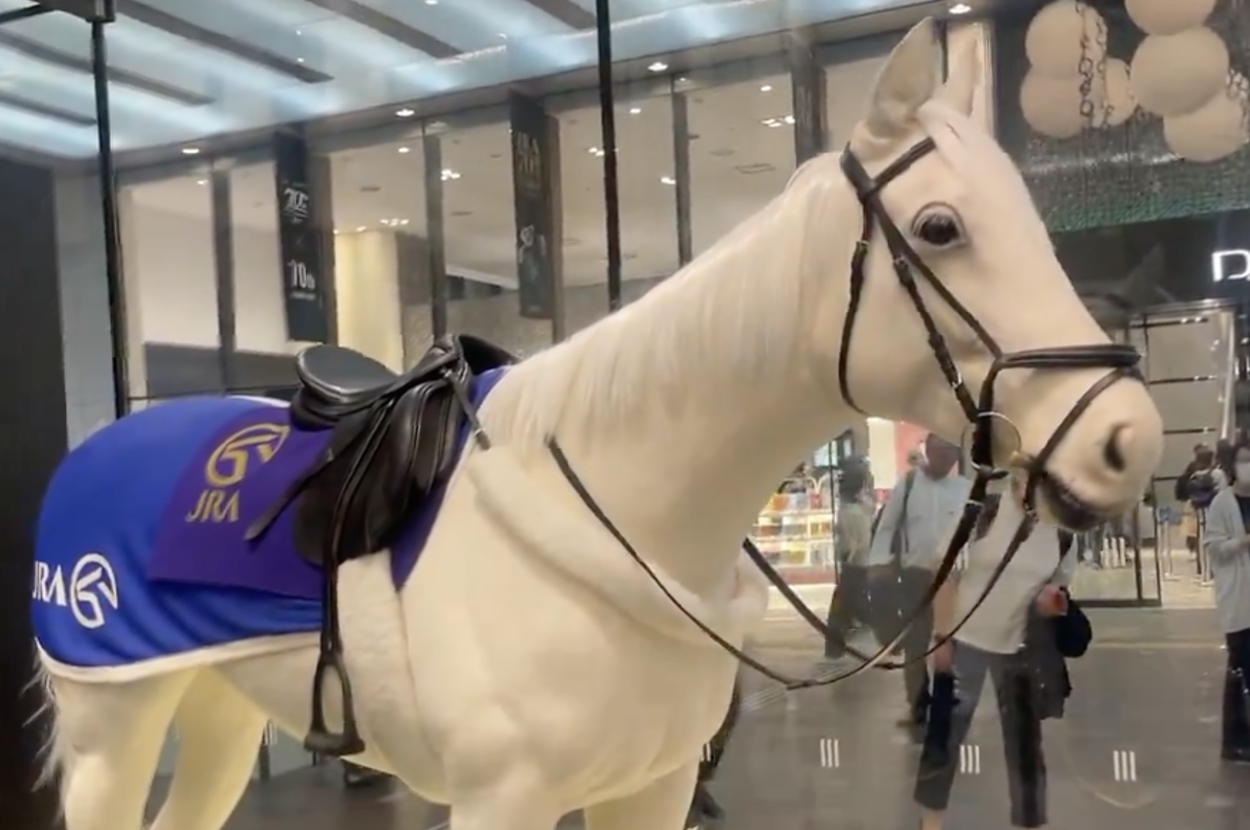 JRAの70周年記念展示の一環としてガラスケース内に置かれた白い馬の彫像。背景に人々が歩いているショッピングモール。