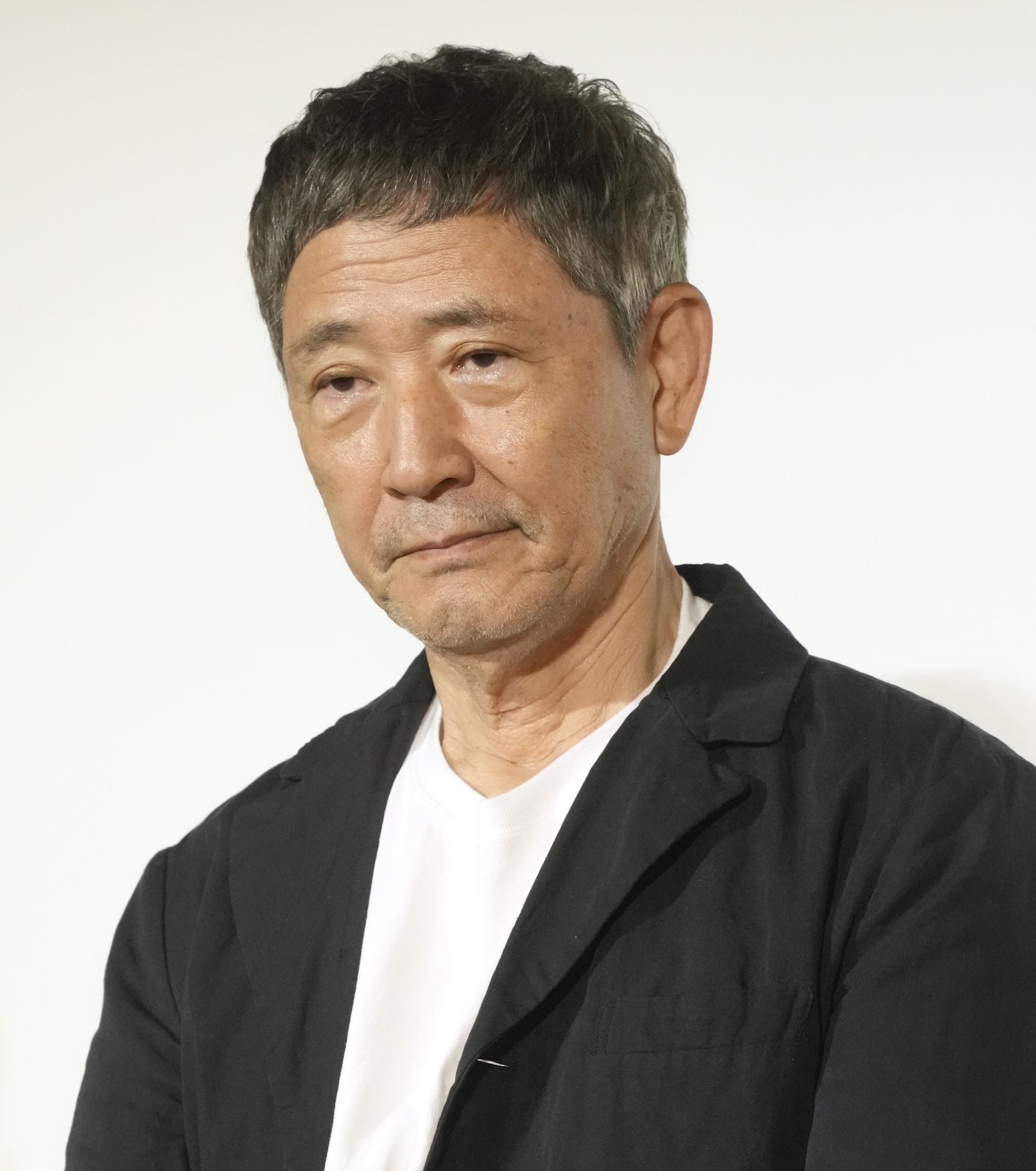 男性俳優の役所広司が、シンプルな黒いジャケットと白いシャツを着て、カメラに向かっている。