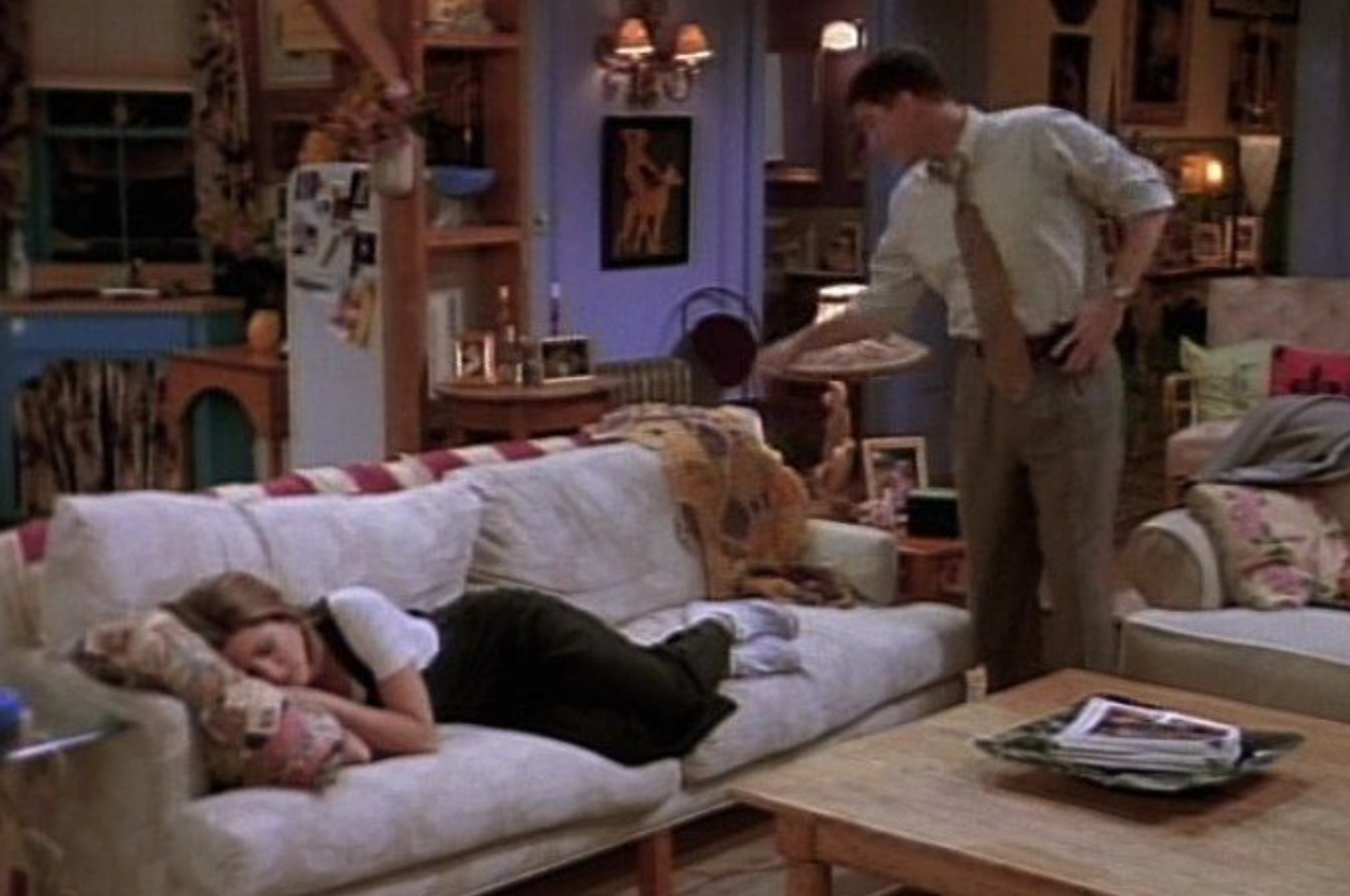 Rachel lies on a sofa with her head on a pillow. Ross stands nearby holding a paper plate. They are in a cozy apartment filled with various items and photos