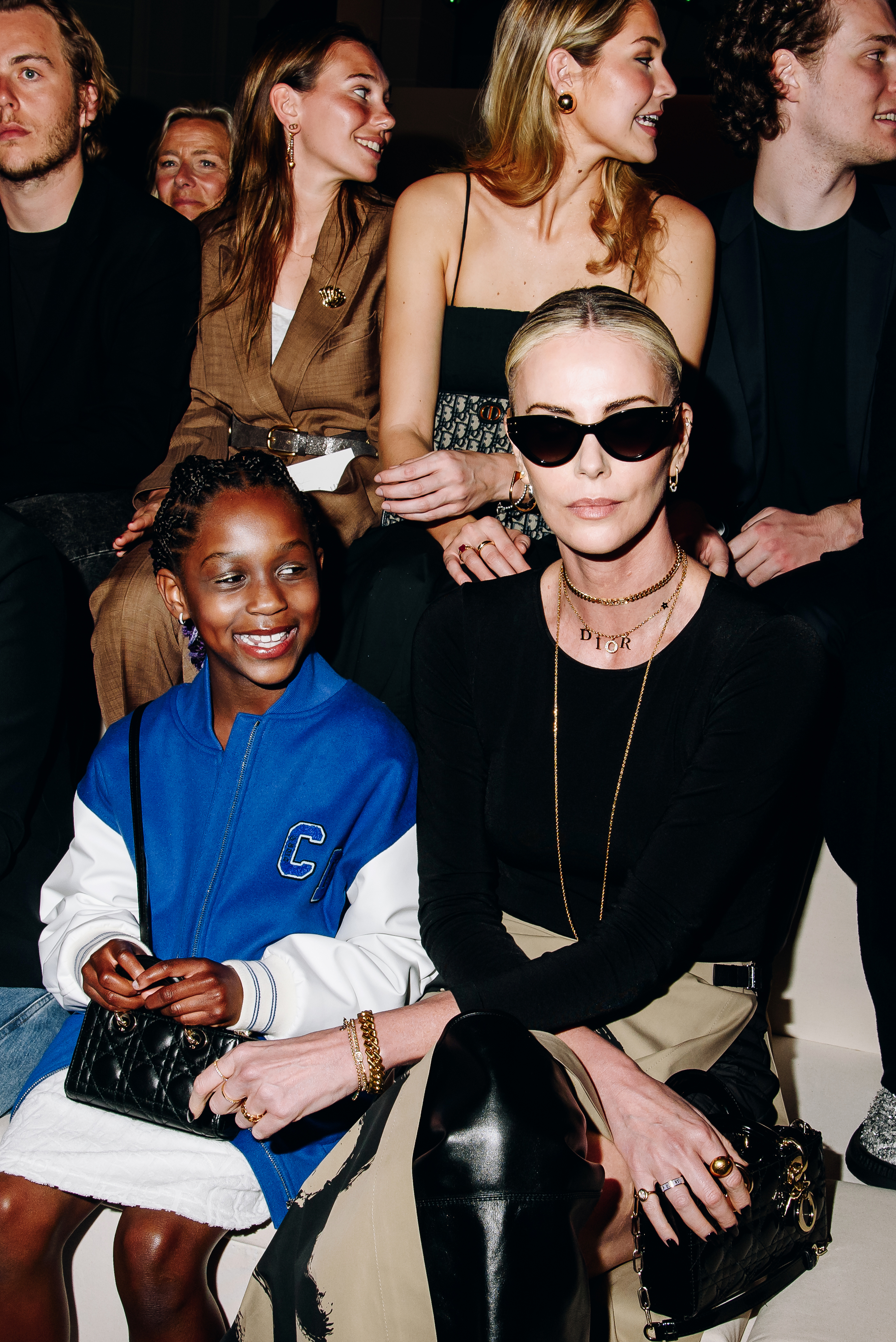 Charlize Theron in a black top and sunglasses sits with her daughter, Jackson Theron, wearing a blue and white jacket, at a fashion event. Others sit behind them