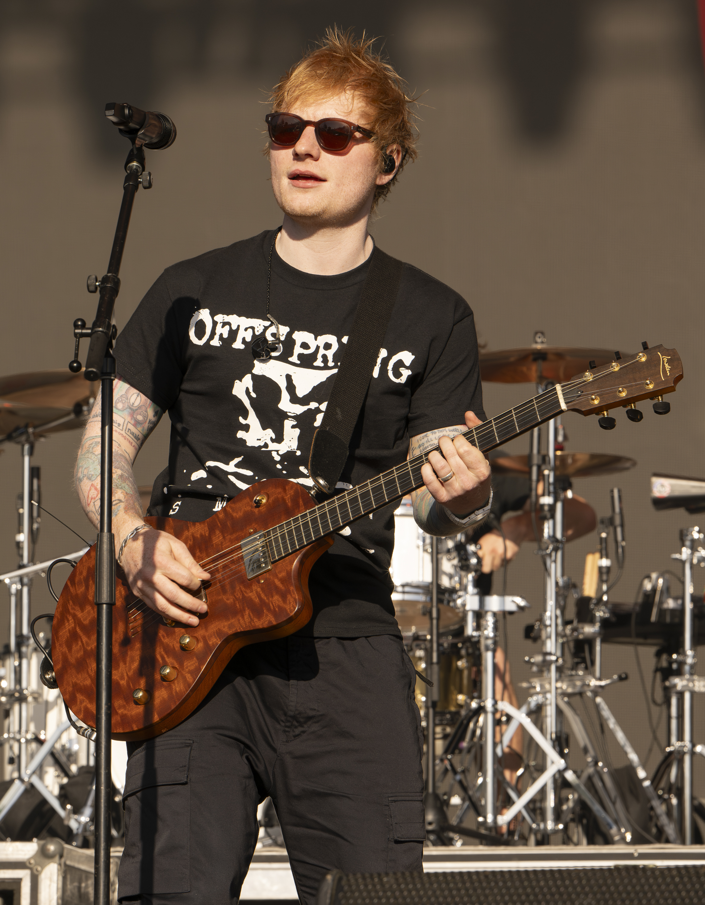 Ed Sheeran performing on stage with a guitar, wearing sunglasses, a black Offspring t-shirt, and dark pants