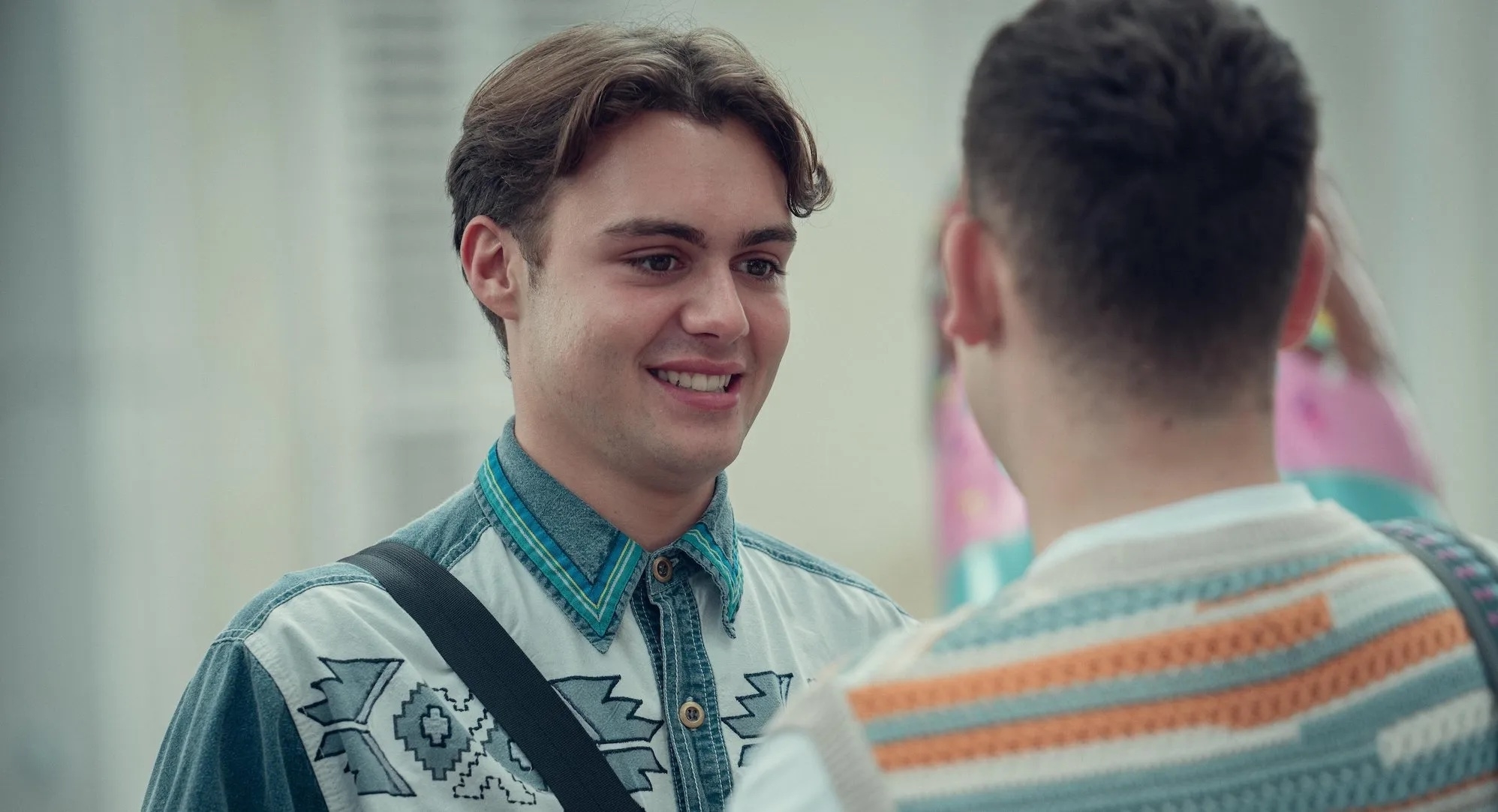 Ncuti Gatwa and Connor Swindells are engaged in conversation. Ncuti wears a denim-style jacket with patterns, while Connor wears a striped knit top