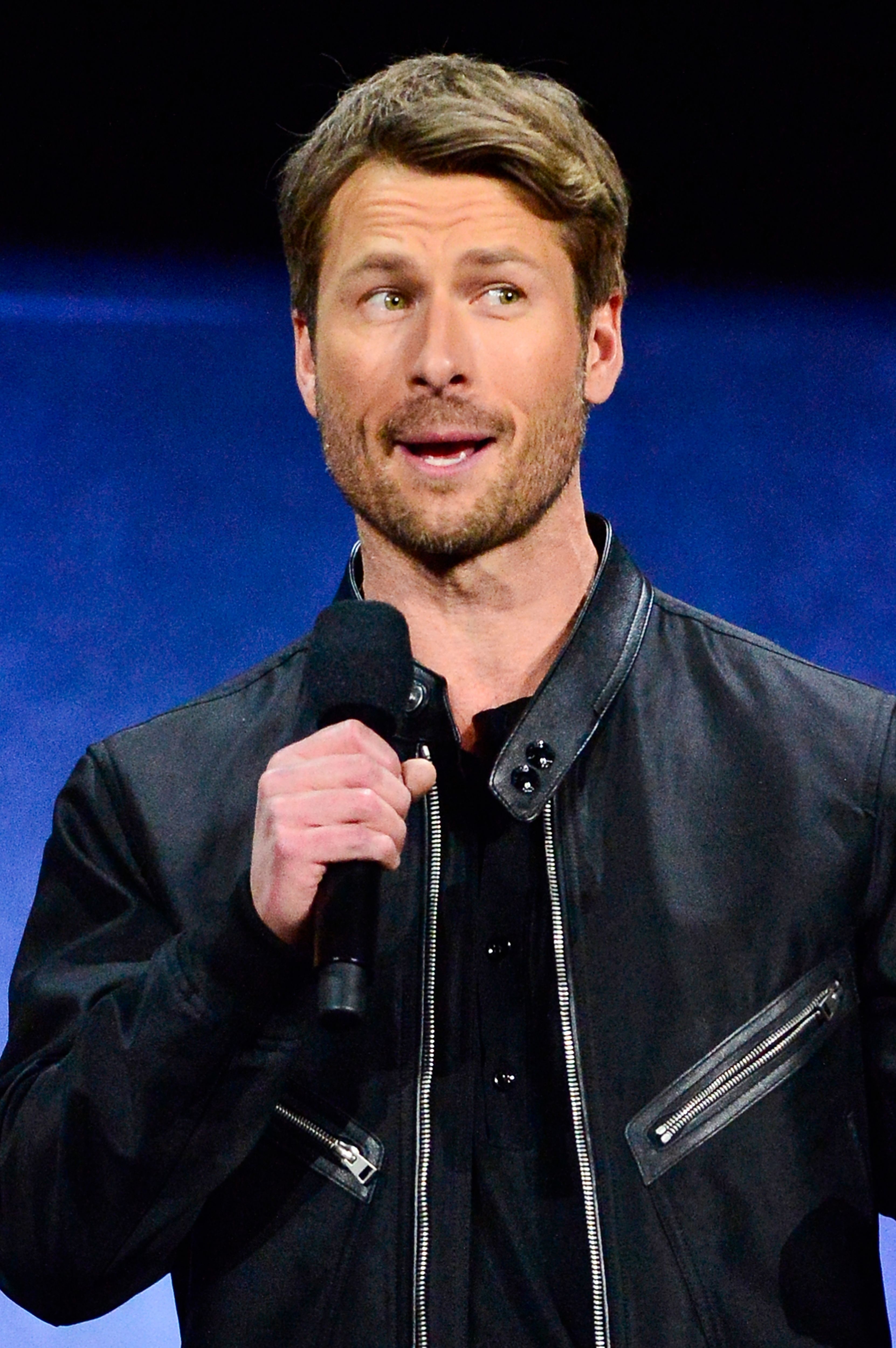 Glen Powell is speaking on stage, holding a microphone, wearing a black jacket with zipper details