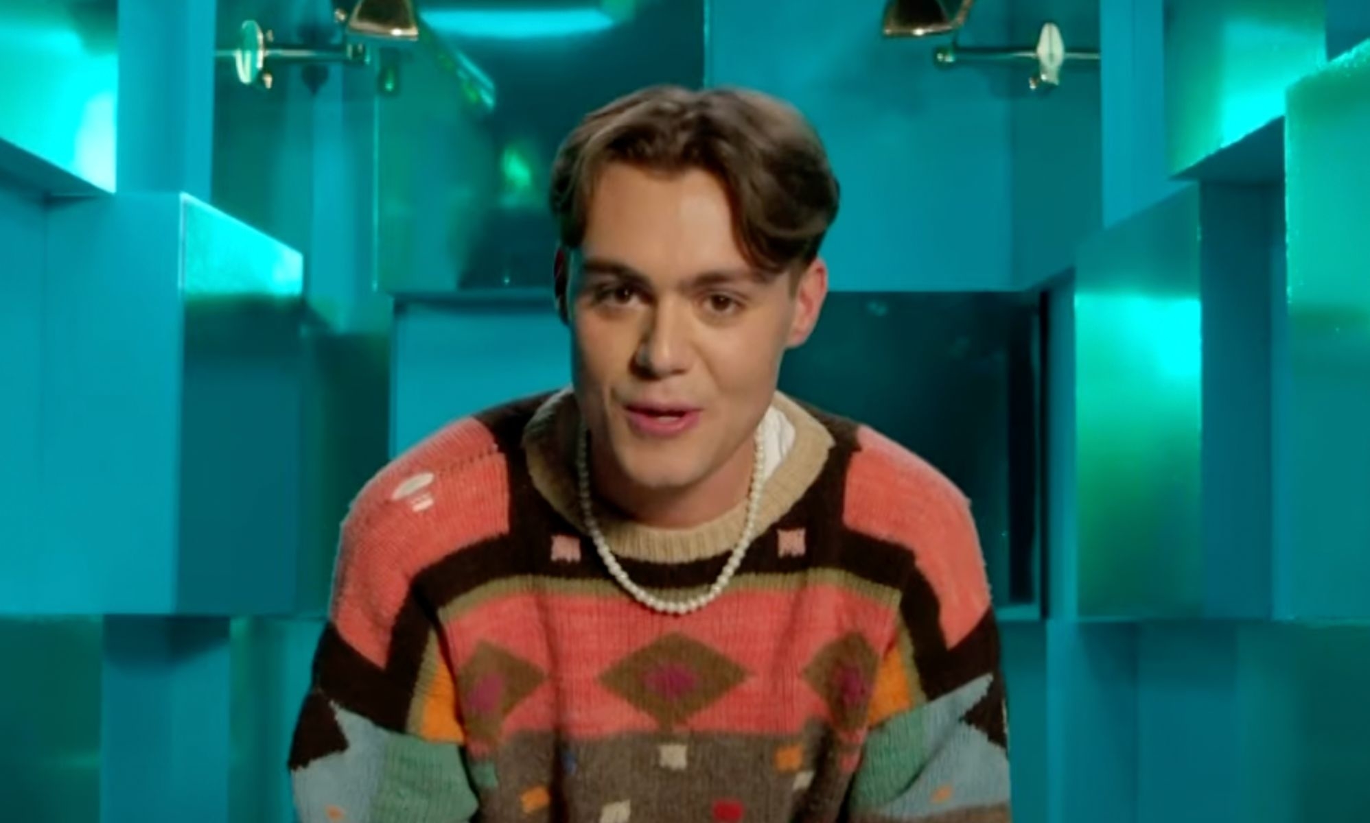 Christian Vance in a colorful geometric-patterned sweater and pearl necklace, speaking in front of a teal background
