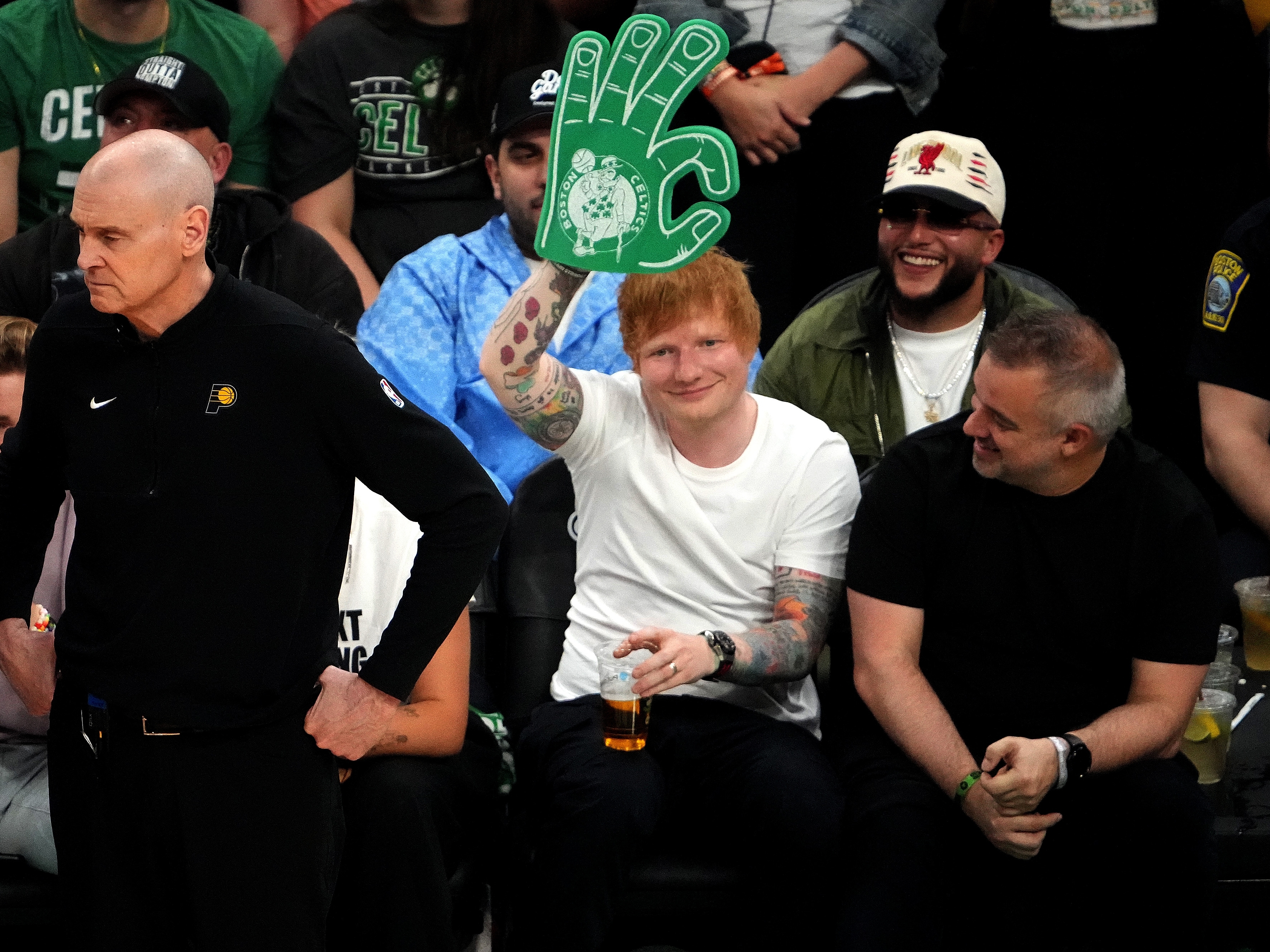 Ed Sheeran holds a foam finger at a basketball game, sitting beside two smiling men. A coach stands in the foreground